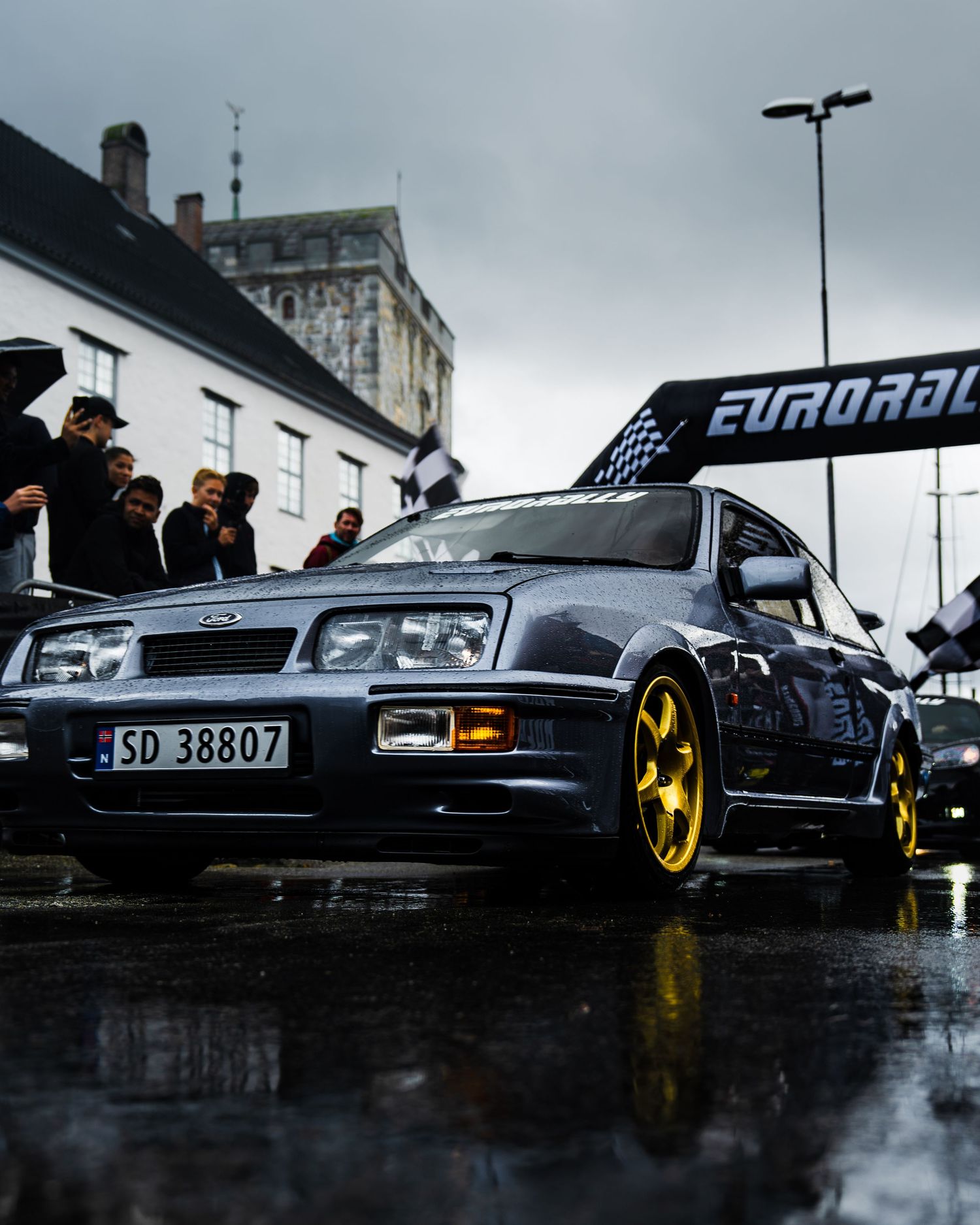 Silver Ford Sierra Cosworth with gold wheels under Eurorally banner on rainy street.