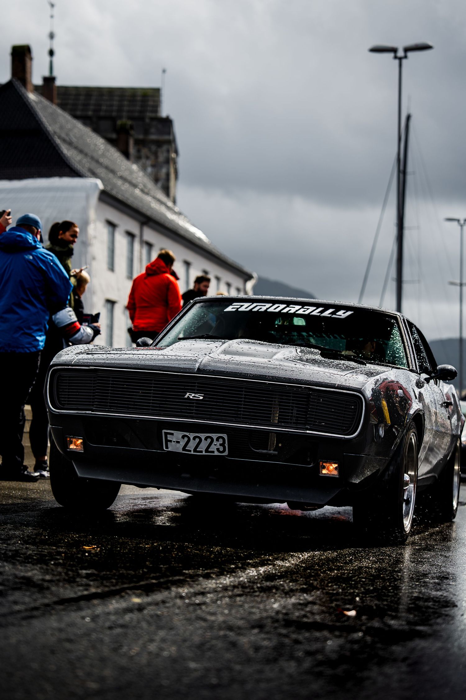 Sleek black vintage car photographed from a low angle on wet pavement during overcast weather.