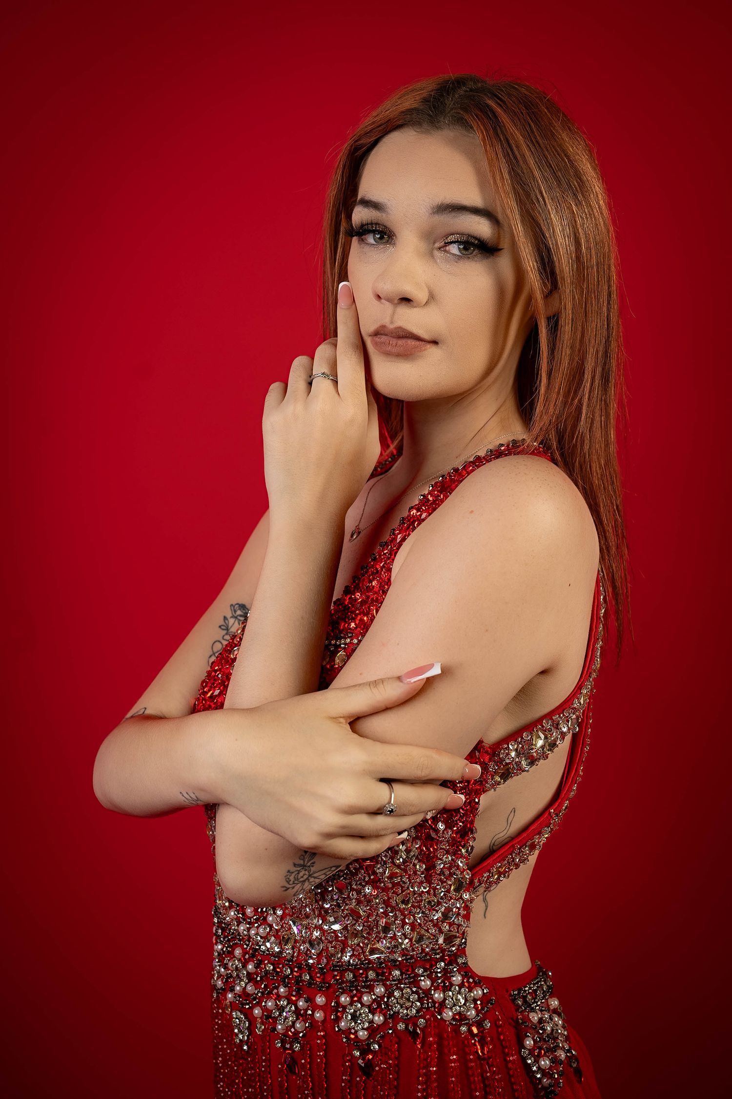 Young woman in red beaded dress poses against red background, showcasing tattoo and elegant jewelry.