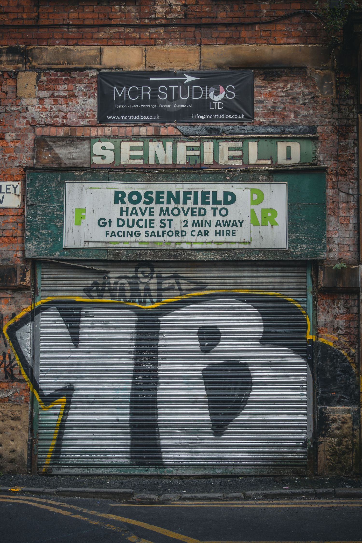 Old brick wall with weathered business signs and metal shutters showing MCR Studios and Senfield signage.