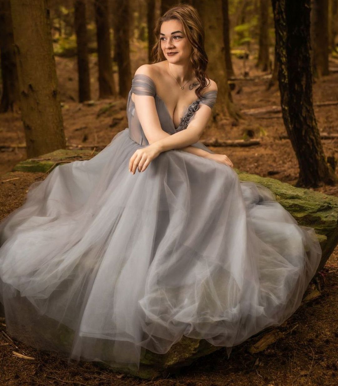 Young woman in elegant gray tulle ball gown poses on mossy rocks in a forest setting.