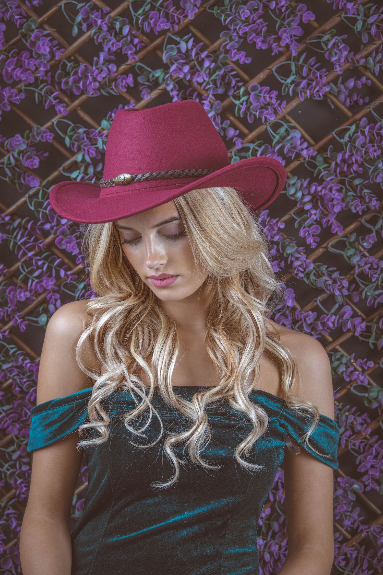 Blonde woman in a burgundy cowboy hat and teal off-shoulder top poses before a purple floral backdrop.