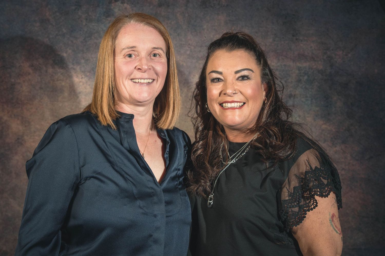 Two smiling women pose together for a portrait photo against a brown studio background.
