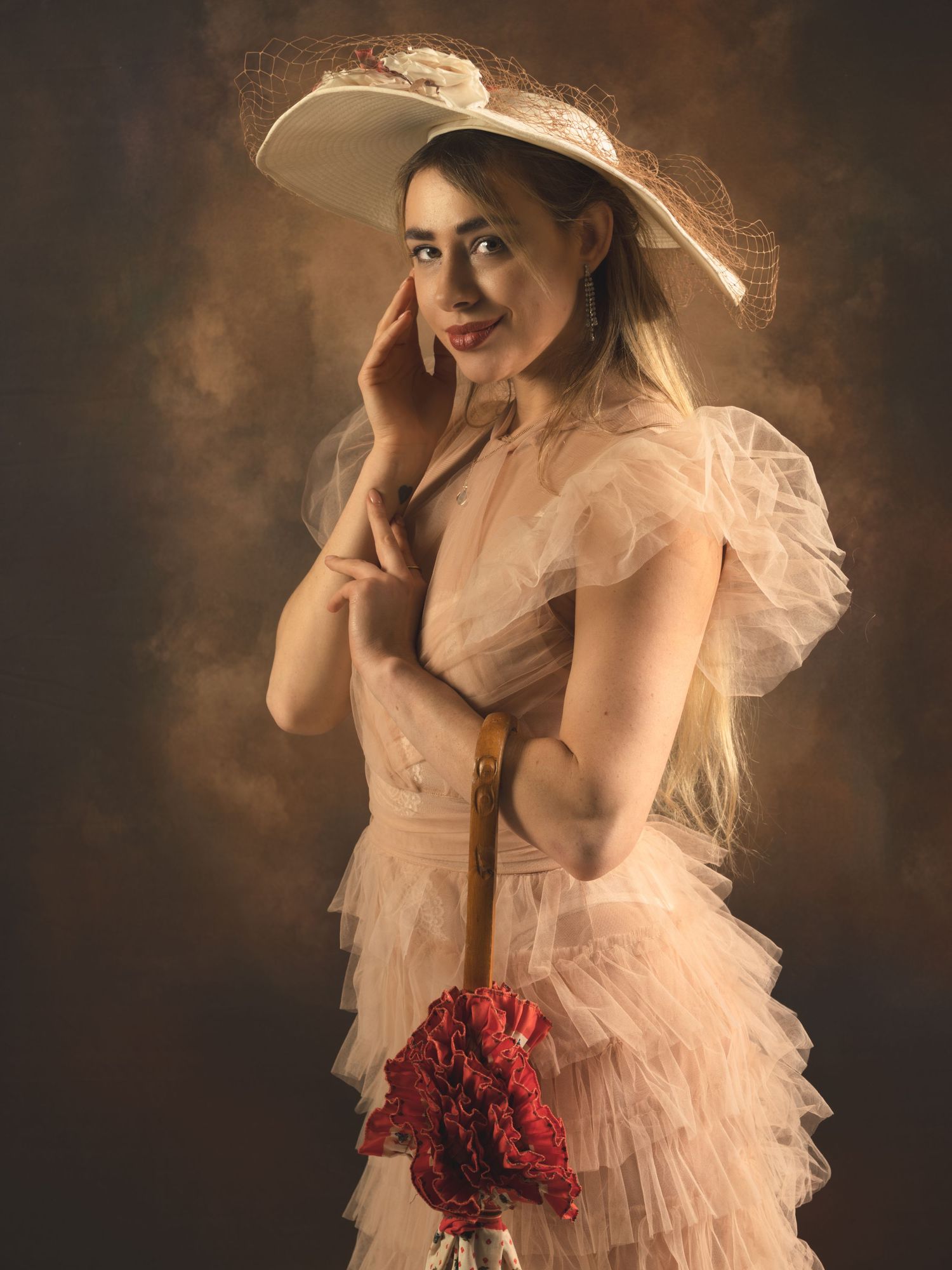 Young woman in vintage pink tulle dress and white hat holding red flowers, posing elegantly against brown backdrop.