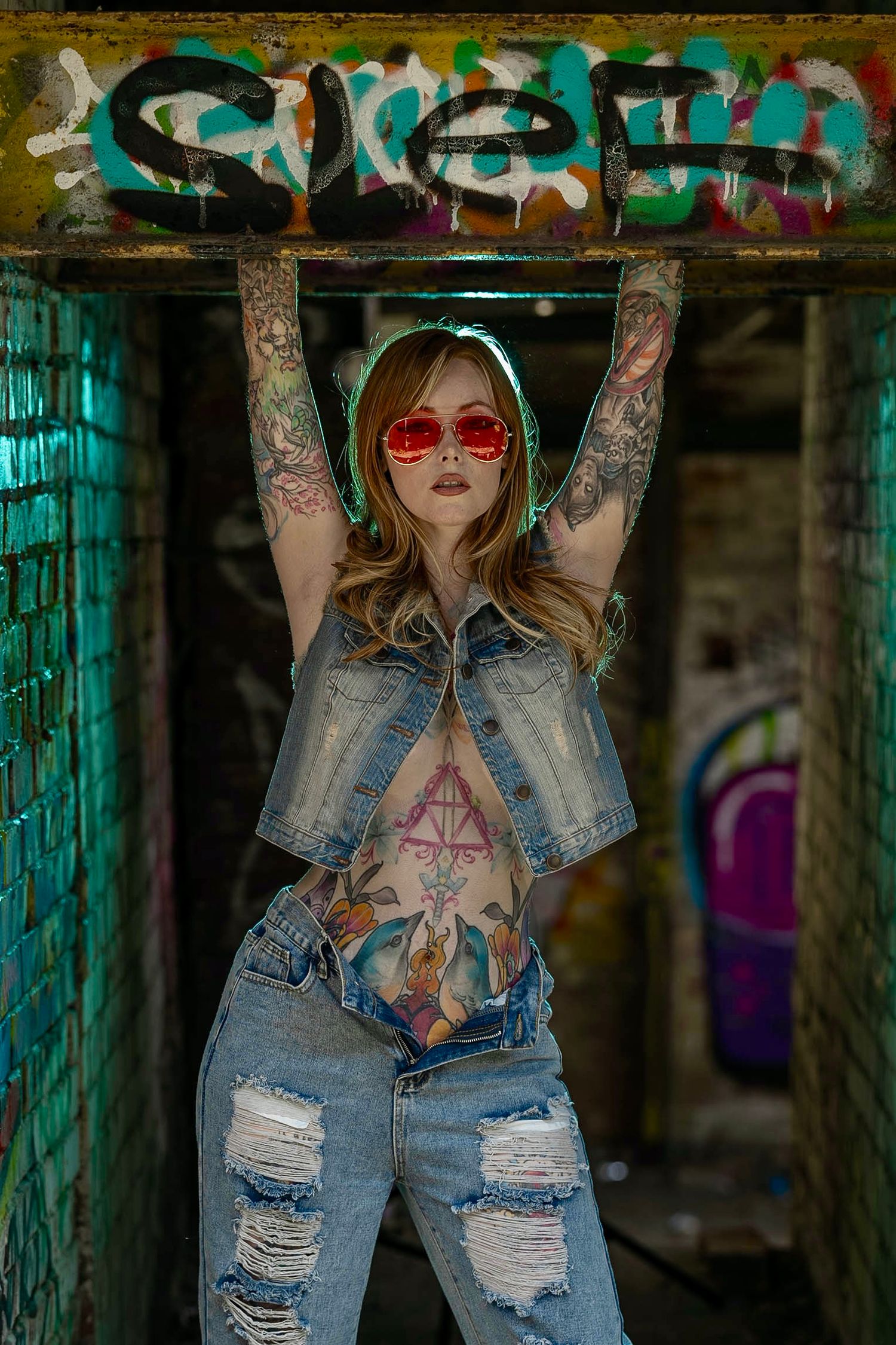 Tattooed woman in ripped denim and red sunglasses poses against graffiti wall in urban setting.