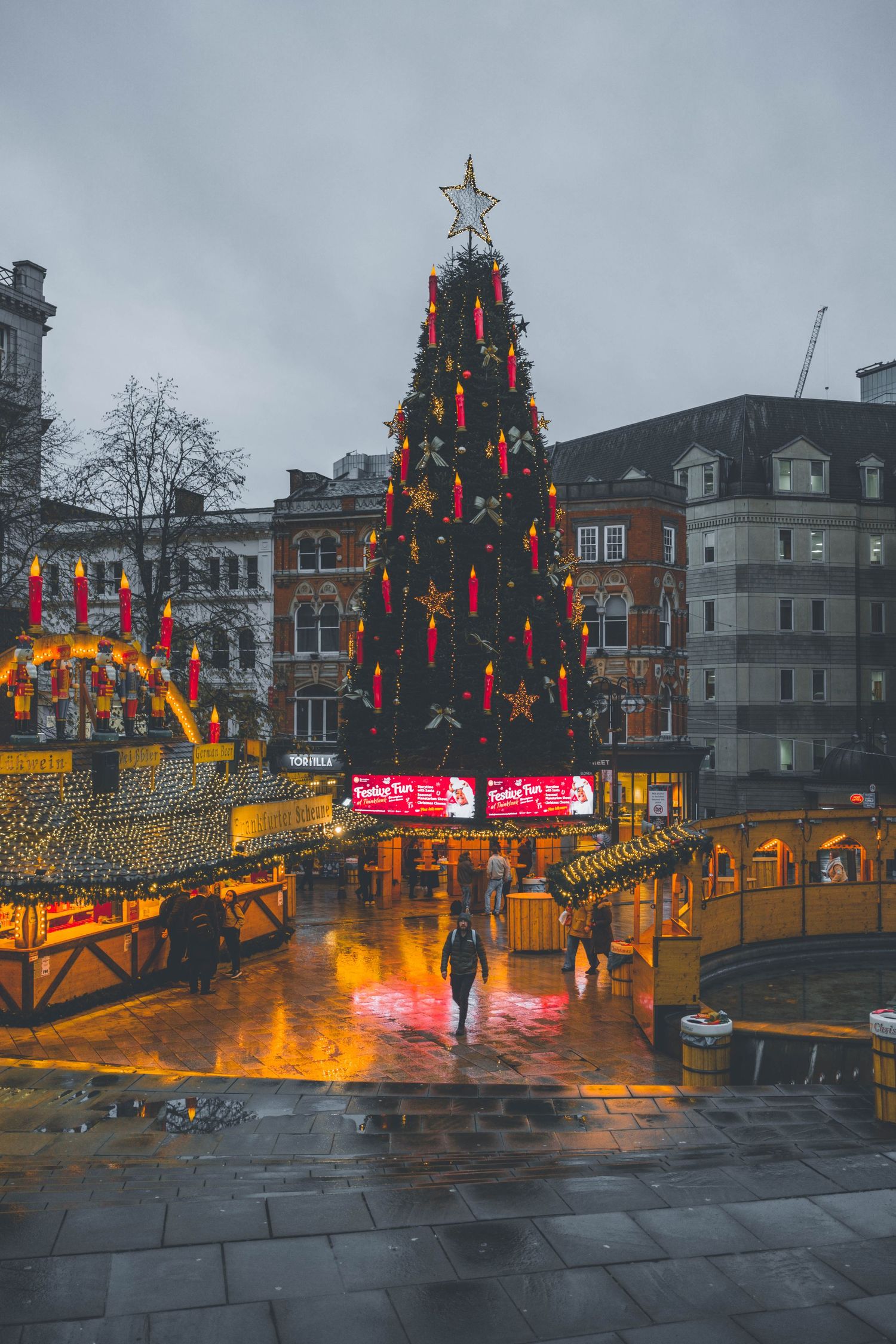 A rainy evening street scene with illuminated Christmas trees and decorations in a town square during the holiday season.
