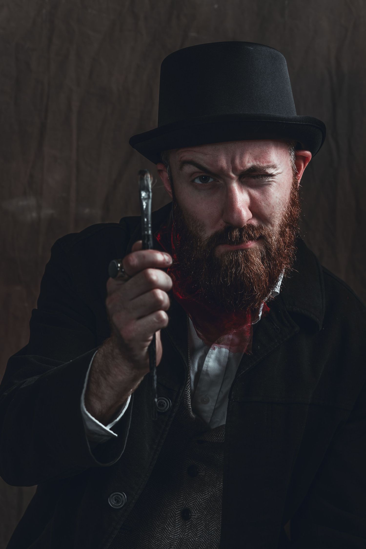 Dark moody portrait of a bearded person in top hat examining something with a magnifying glass.