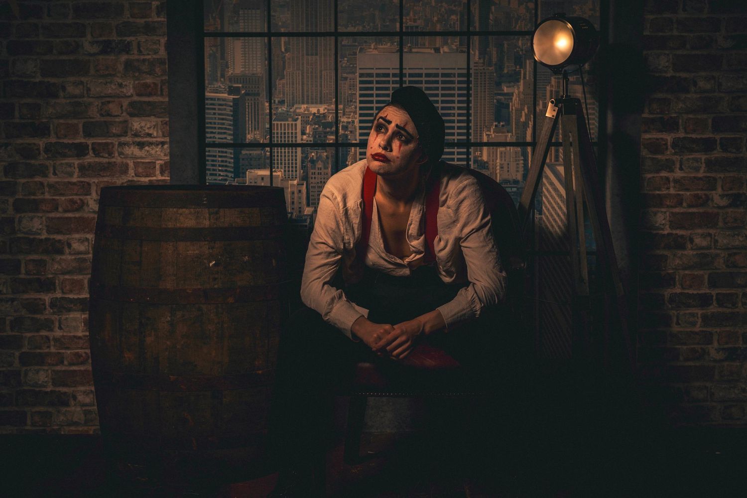 A brooding man in suspenders sits pensively beside a wooden barrel in a dimly lit, rustic brick room.