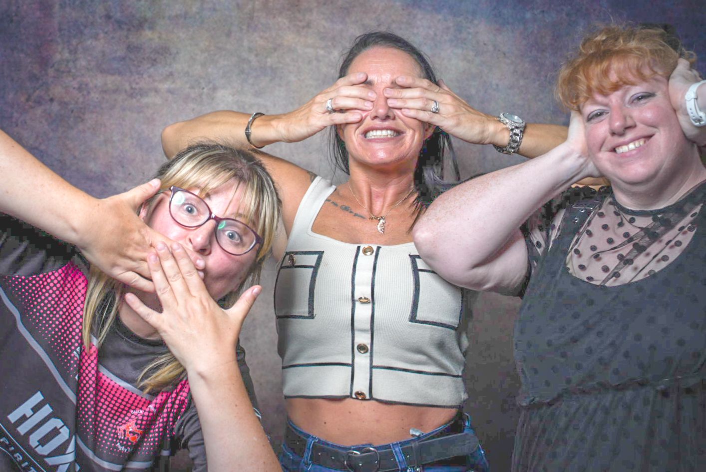 Three women posing playfully, covering eyes and making funny faces in a photo booth setting.