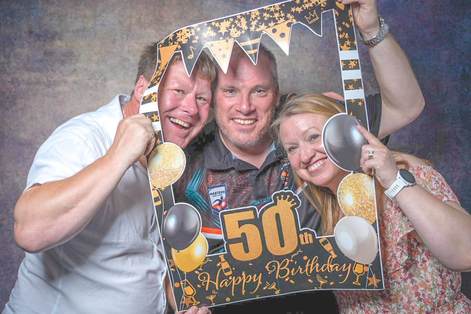 Three people pose with a decorative 50th Happy Birthday photo booth frame, smiling and holding spoons as props.