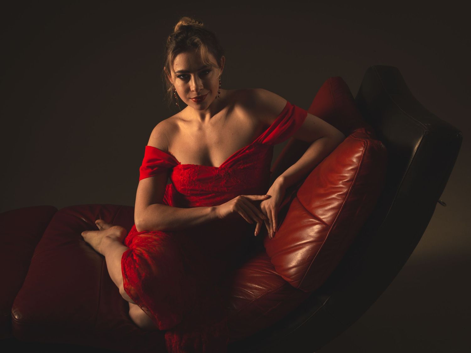 A woman in a red off-shoulder gown poses elegantly on a red leather chair in dramatic studio lighting.