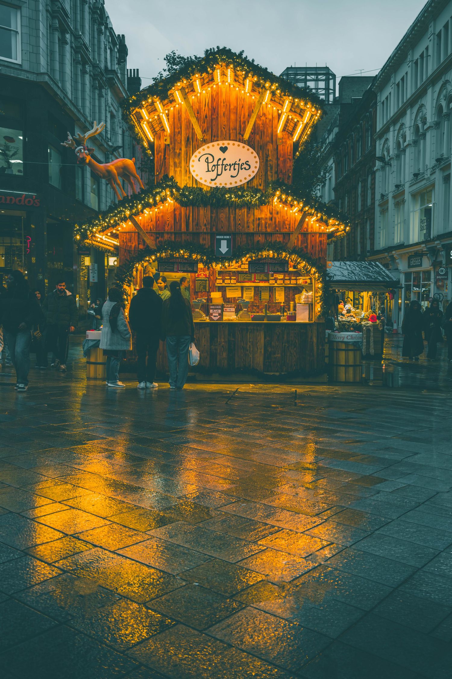 Illuminated Christmas market entrance glows warmly on wet cobblestone street during evening rain in European city.