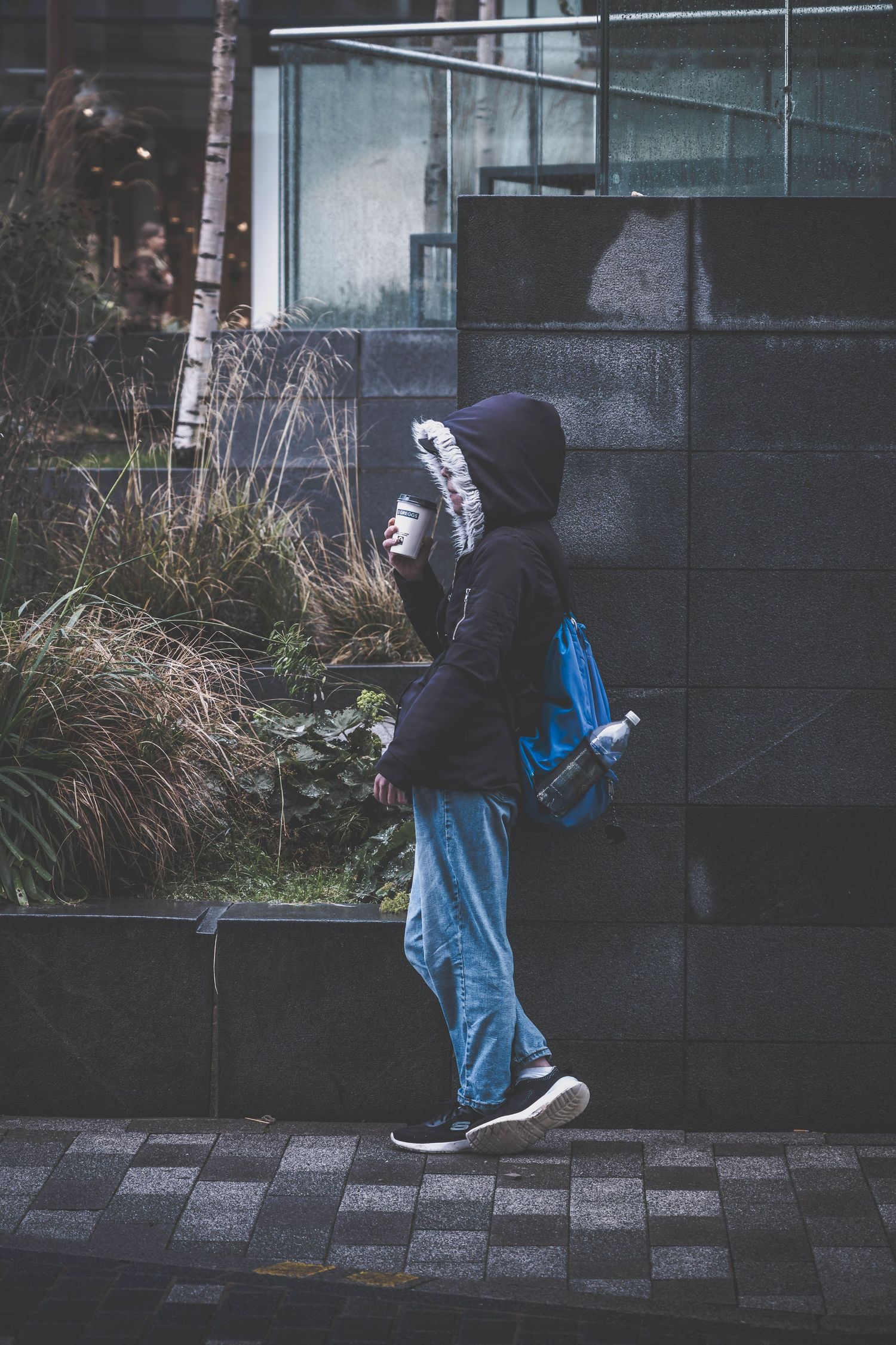 A person in a dark hoodie and jeans stands against a modern glass building in moody urban lighting.