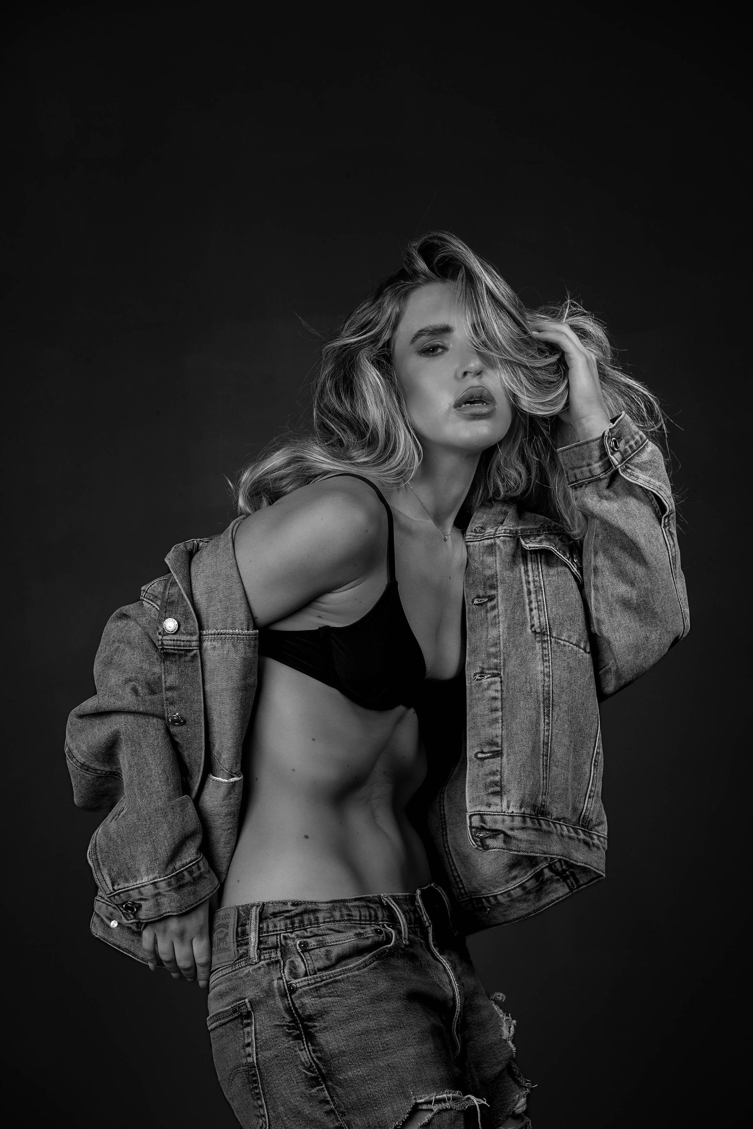 Black and white photo of a blonde woman in a denim jacket and crop top posing confidently in a studio.