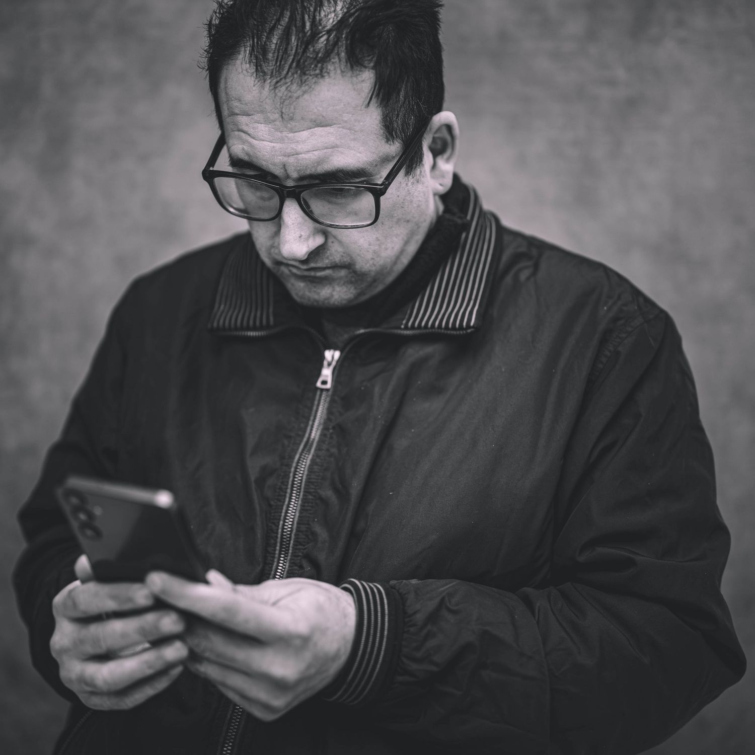 Black and white photo of a middle-aged man wearing glasses and a jacket, looking down at his smartphone.