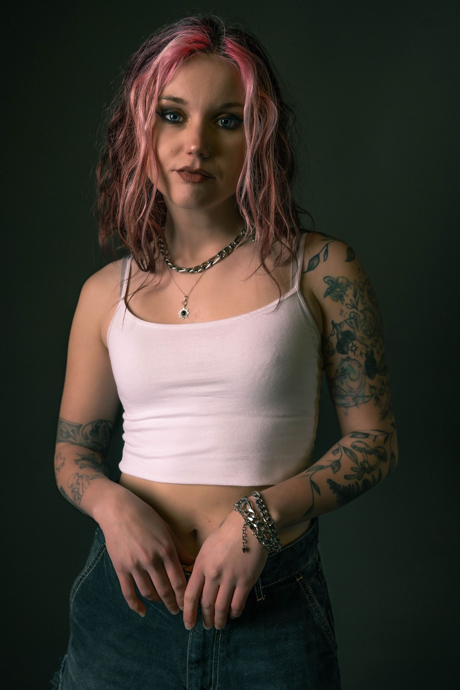Young woman with pink hair and tattoos wearing a white crop top and jeans poses against a dark background.