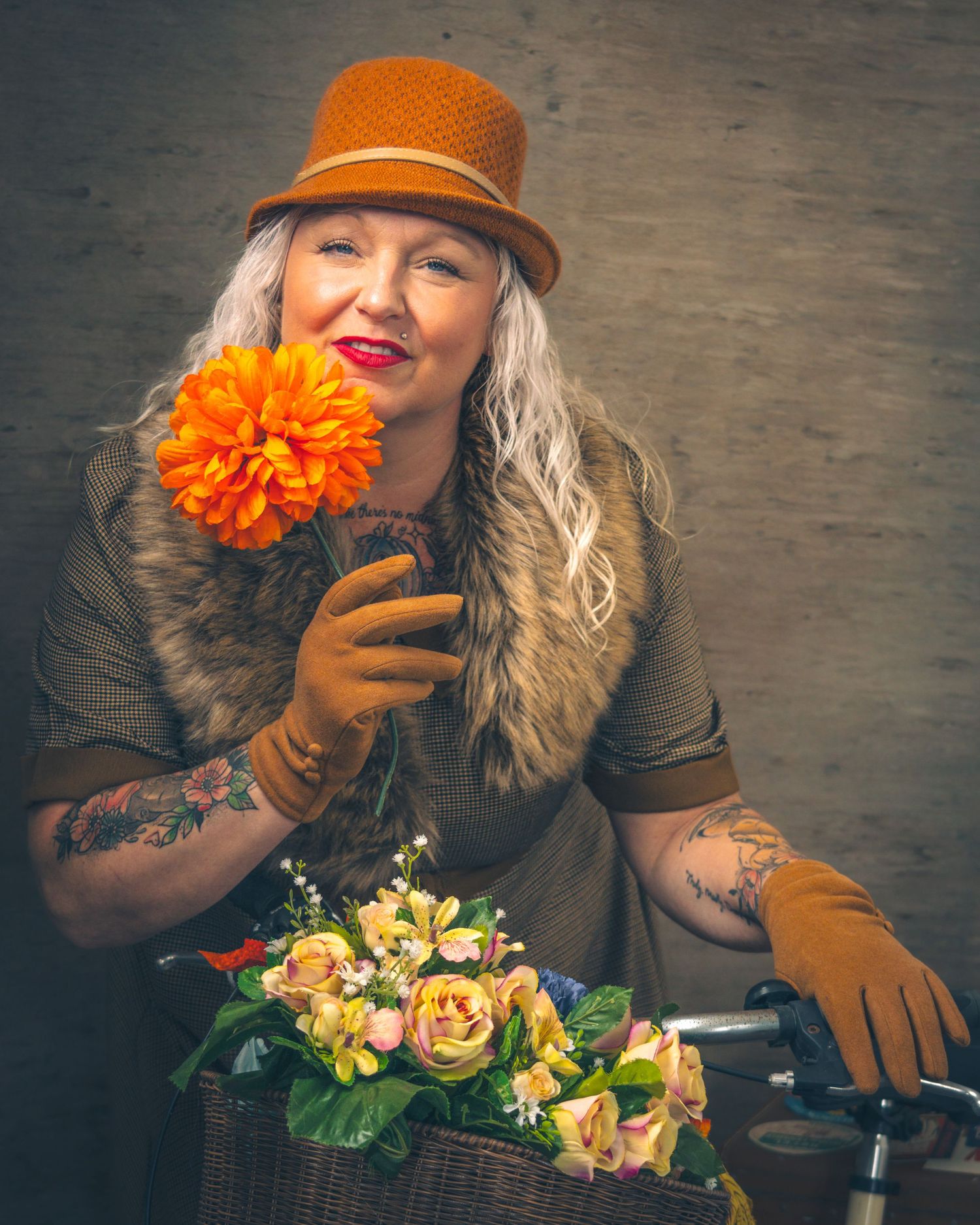 A person in an orange hat holds a bright marigold flower while posing with a bicycle basket full of yellow roses.