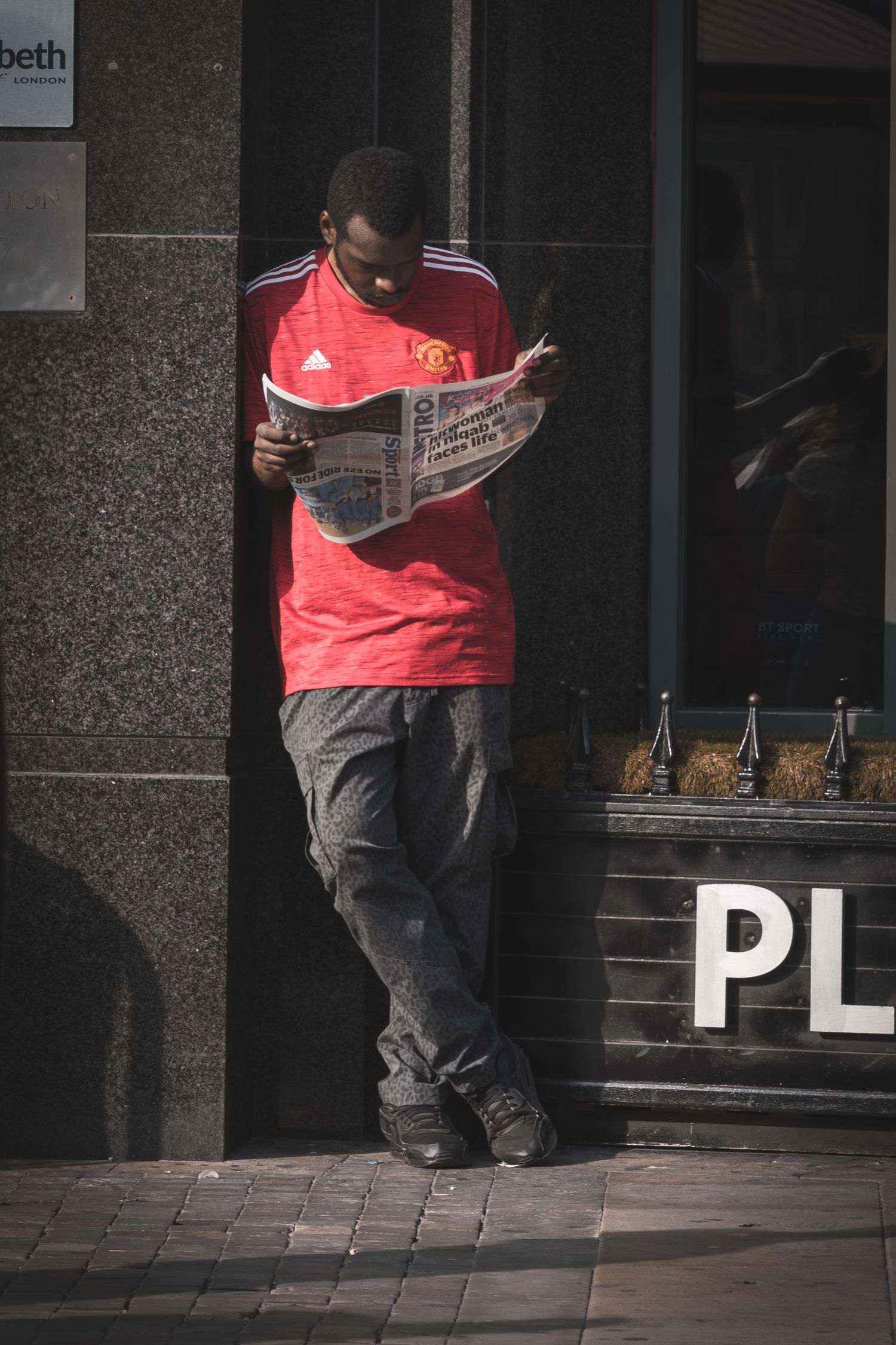 Person in Manchester United jersey and dark pants sits reading on brick pavement outside a building.