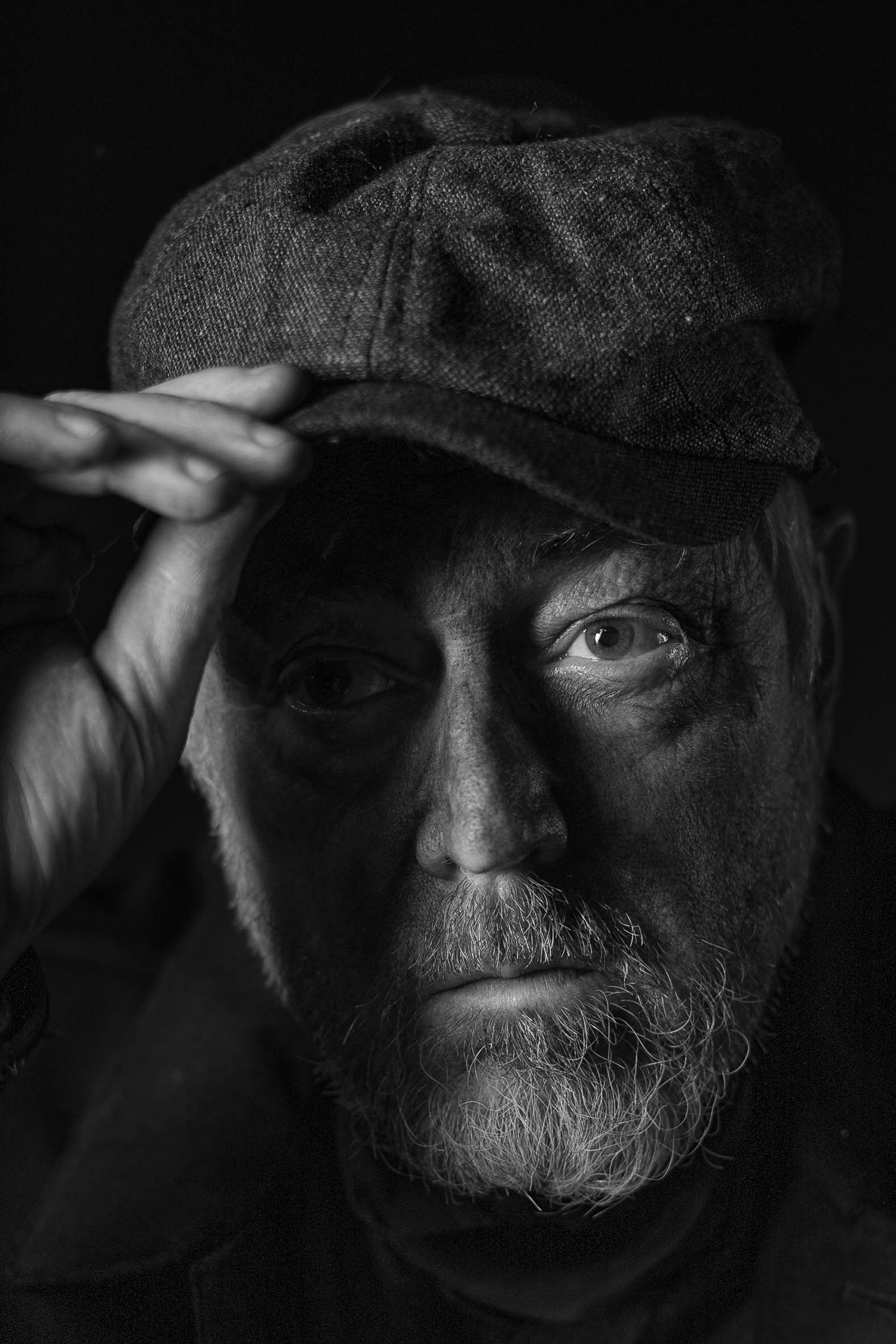 Black and white portrait of an elderly bearded man in a flat cap, gazing intensely at the camera.