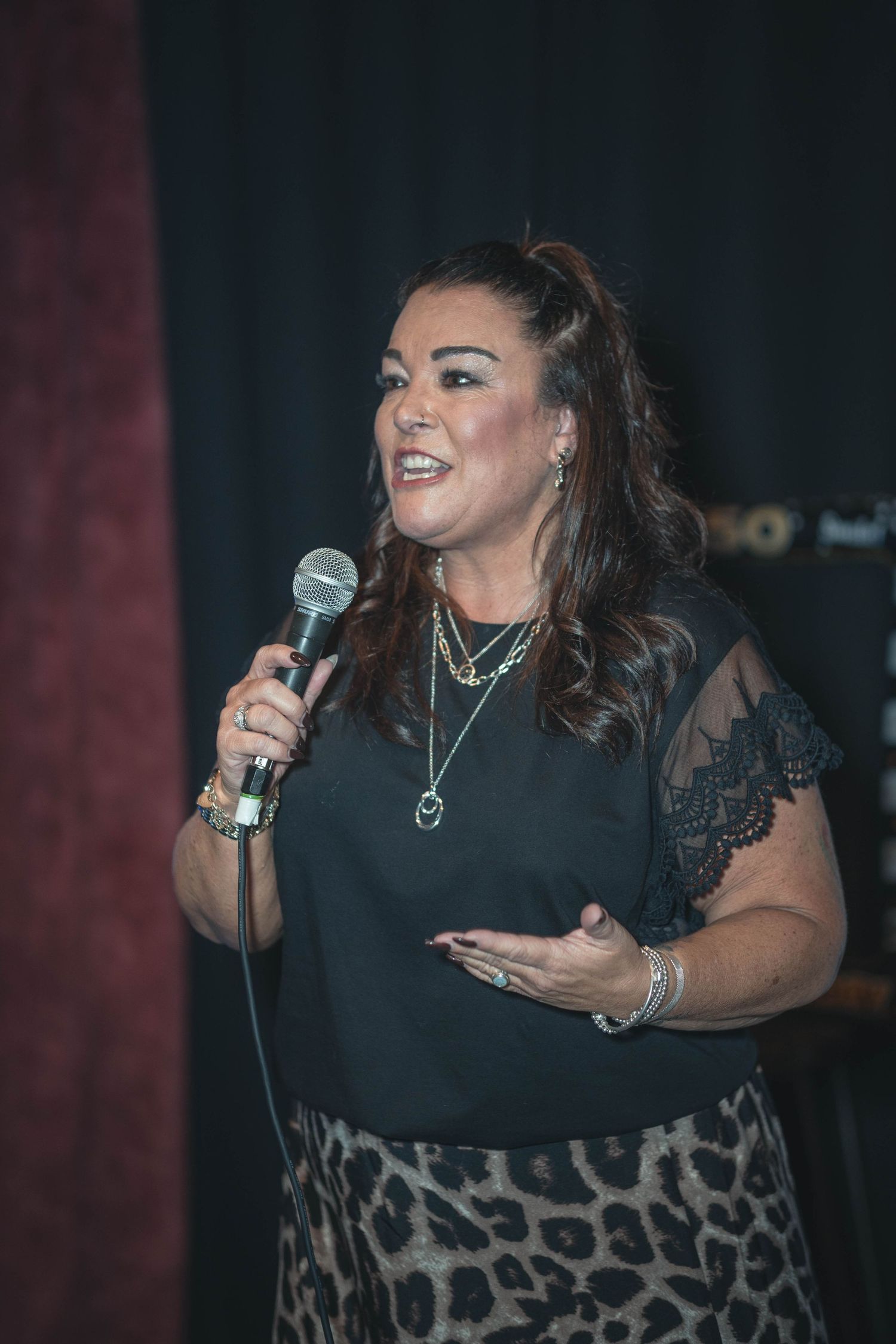 Woman in black lace top holding microphone performing on stage wearing layered necklaces and leopard print skirt.