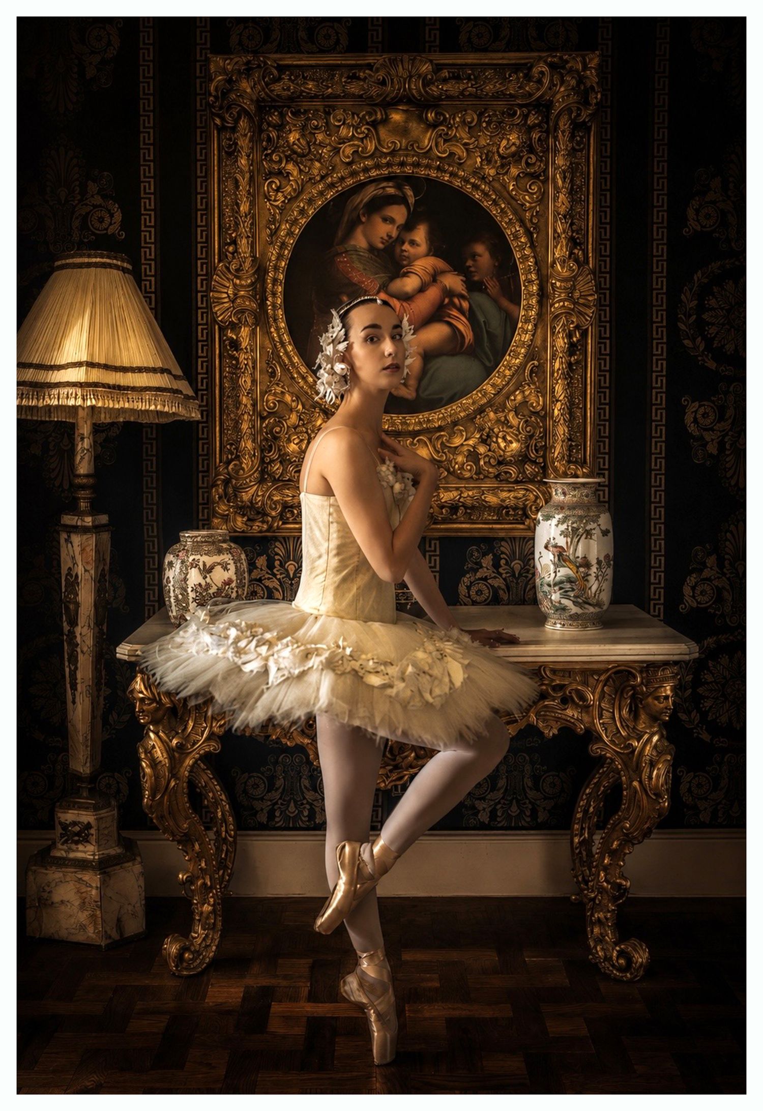 A ballerina in a gold tutu poses en pointe before an ornate gilded table and classical painting in an elegant room.