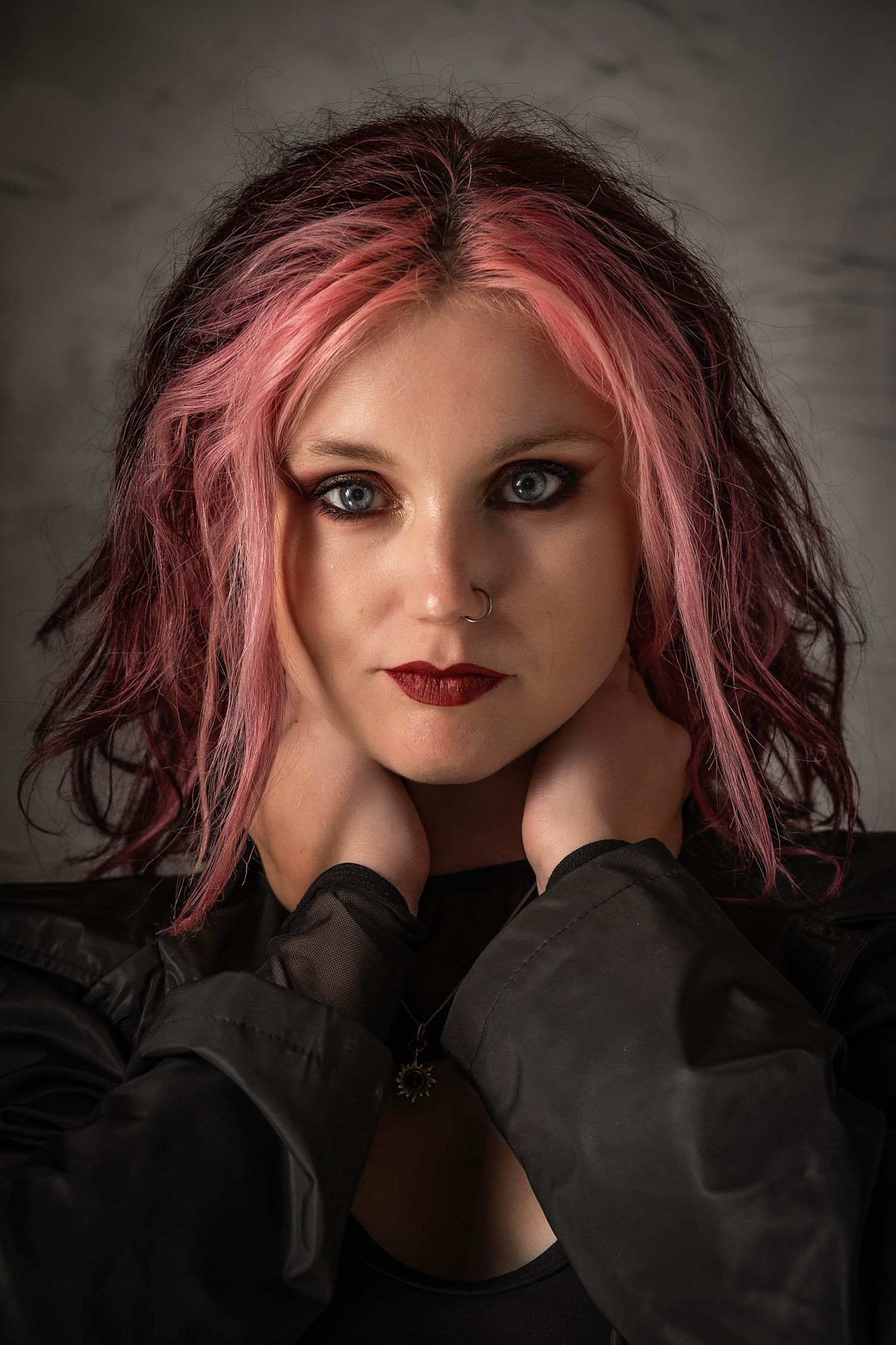 Portrait of a woman with pink-dyed hair, blue eyes, dark red lips, and black outfit posing against a gray background.
