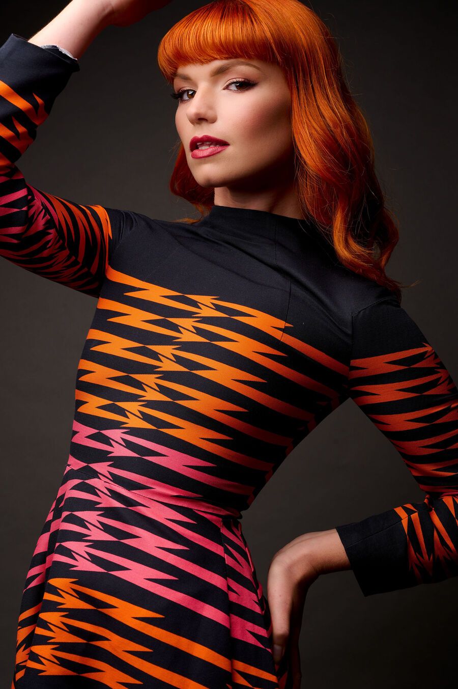 Woman with red hair in black, orange, and pink zigzag patterned long-sleeve dress posing against dark background.