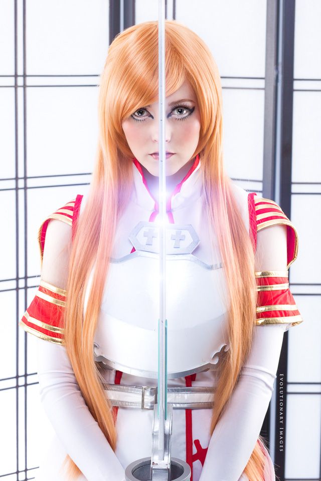 Cosplayer in white and red armor costume with long orange hair holding a glowing sword, anime character cosplay.