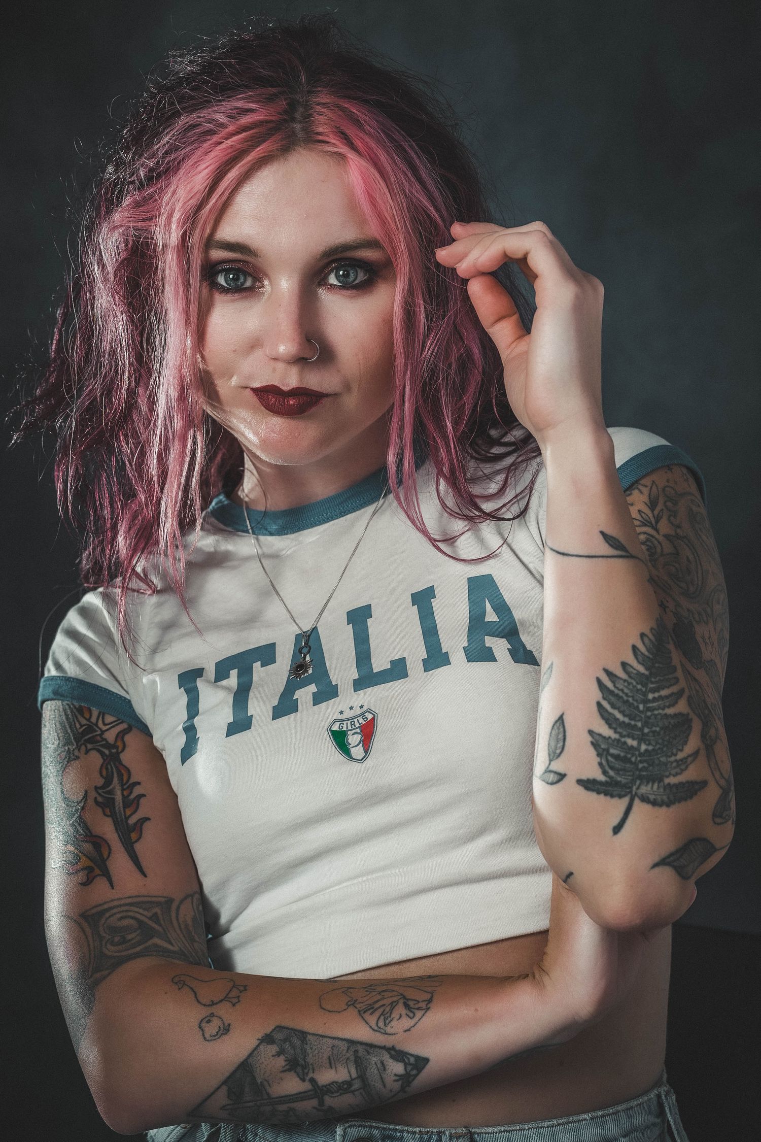 Young woman with pink hair and tattoos wearing an Italia shirt poses confidently against a dark background.