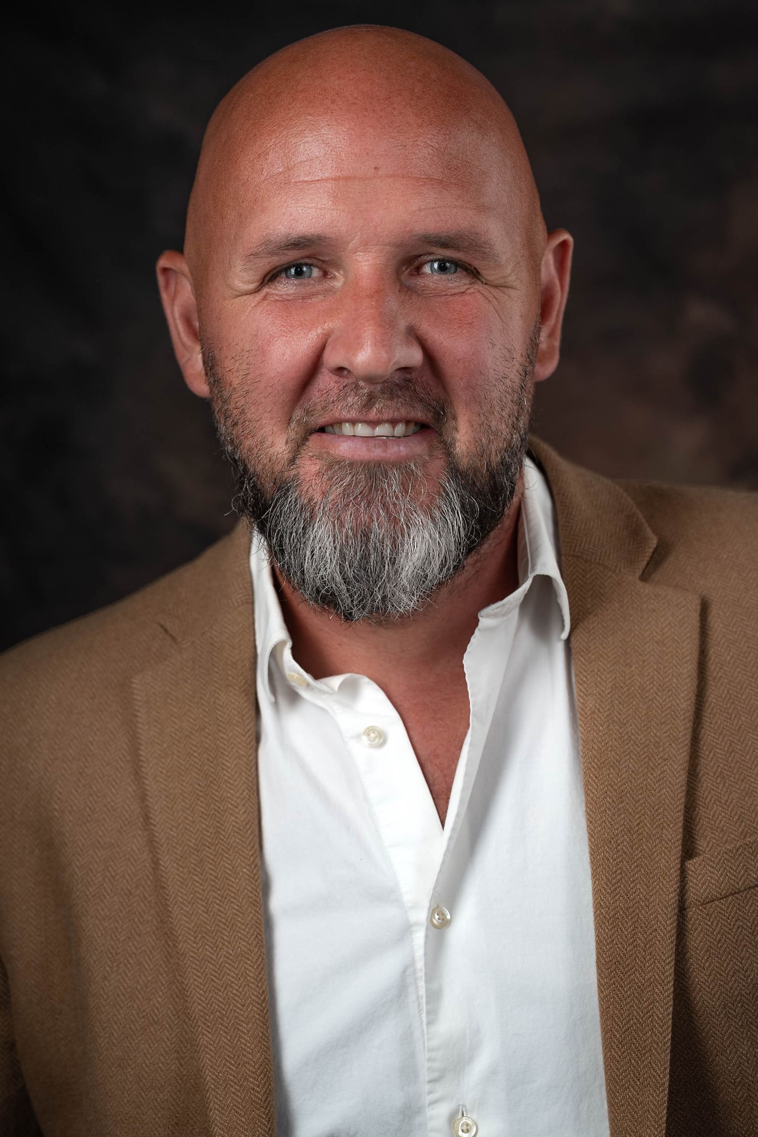 Smiling bald man with a gray beard wearing a white shirt and tan blazer poses for a professional headshot.