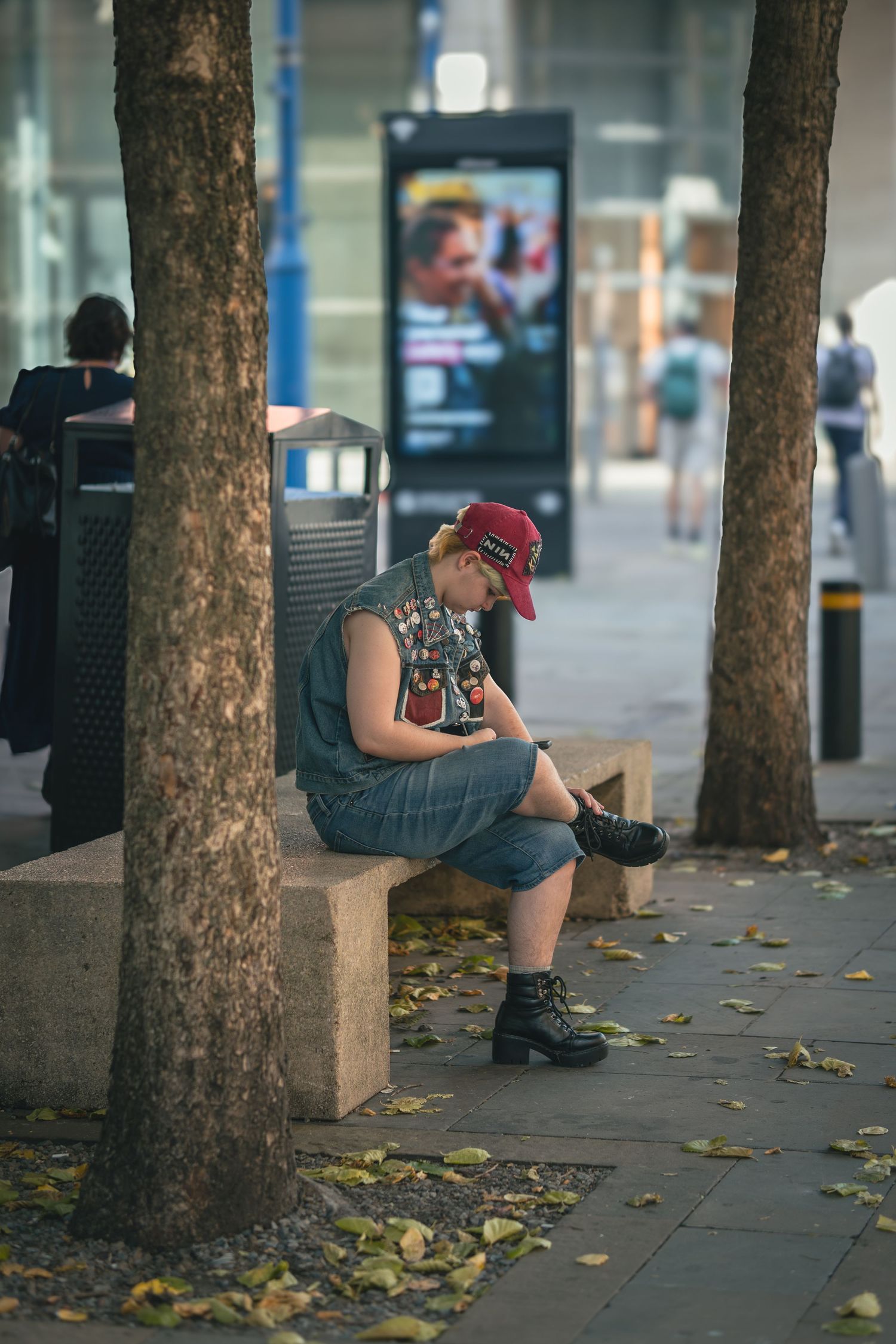 Someone sits on a low ledge against a row of trees on a city sidewalk, wearing casual clothes and a red cap.