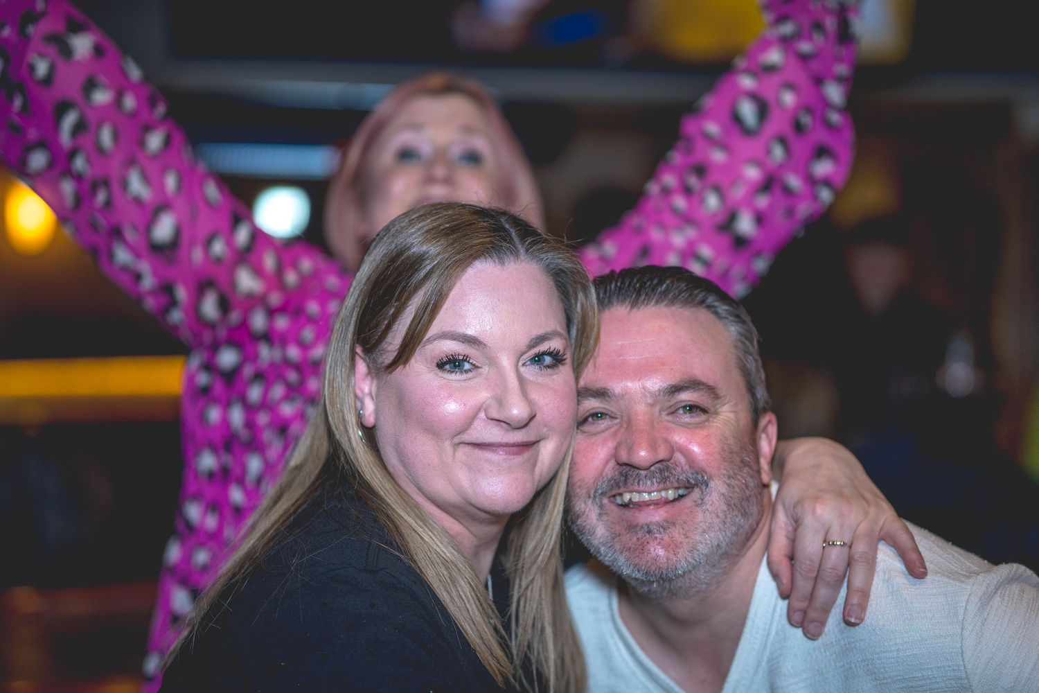 A smiling couple poses together at a bar while a woman in pink raises her arms behind them.
