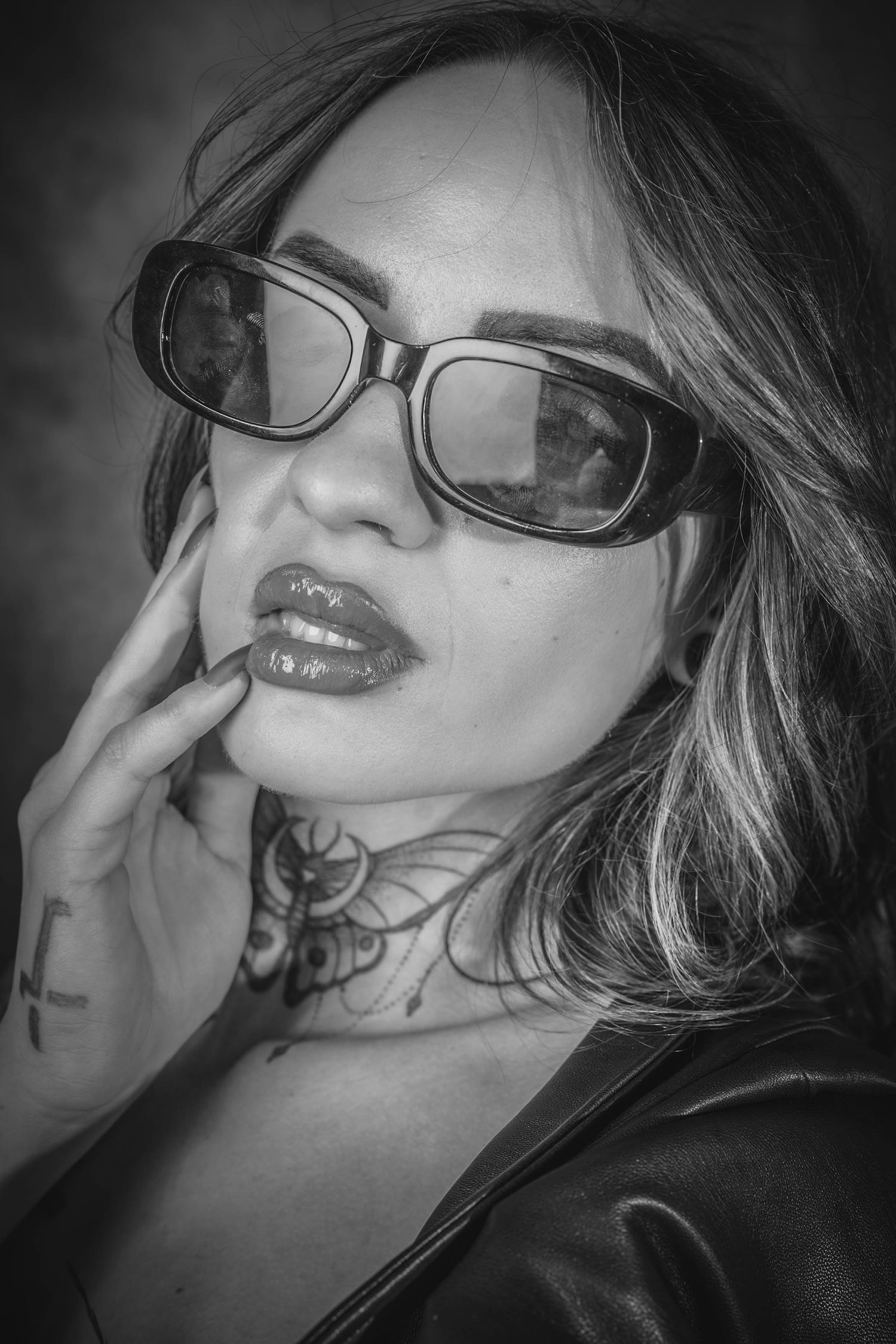 Black and white portrait of a tattooed woman wearing retro sunglasses and leather jacket, touching her face.