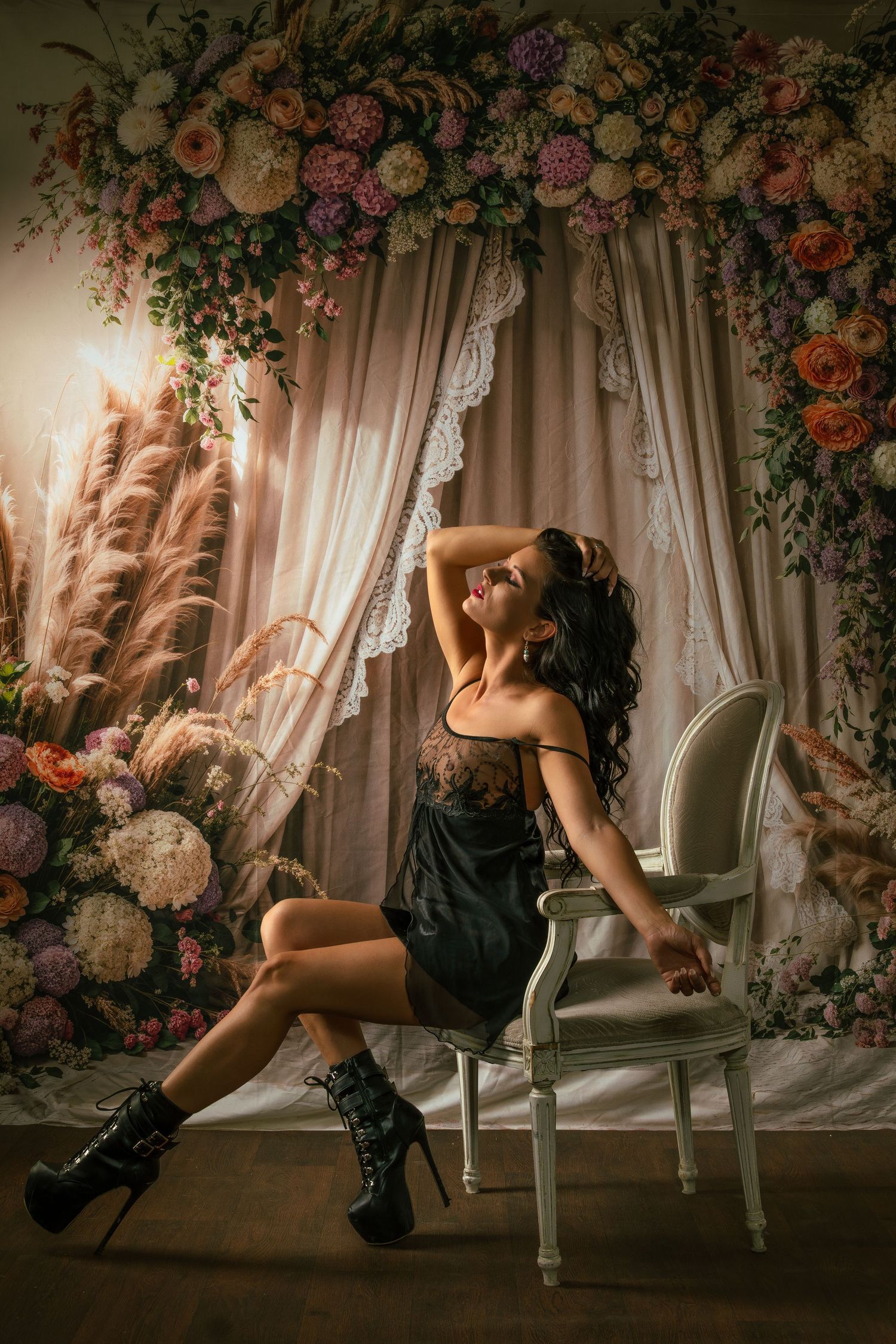 Woman in black dress and heeled boots poses on white chair before floral arch backdrop.