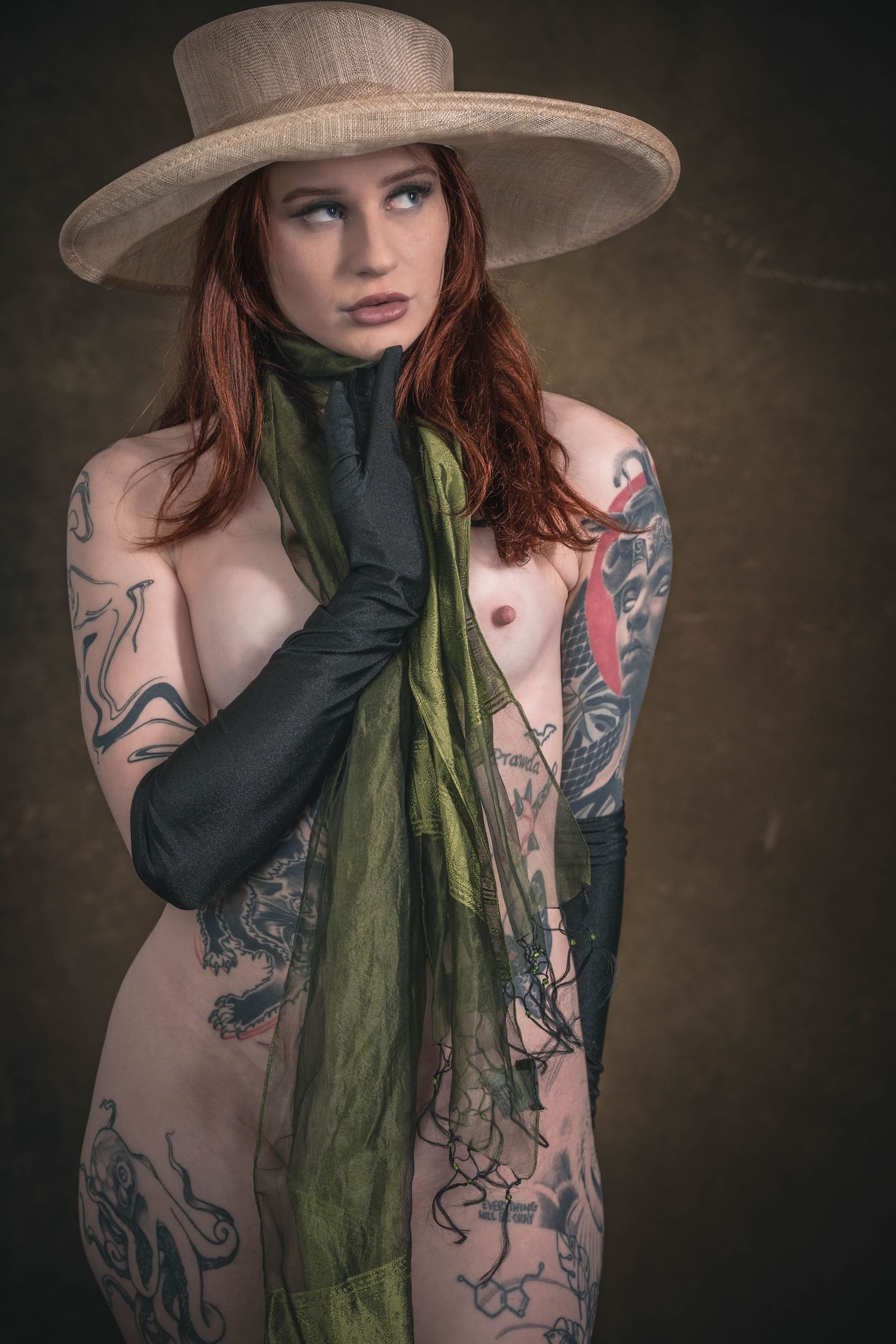 Tattooed redhead woman wearing a wide-brim hat and black gloves holding a green scarf in an artistic portrait.