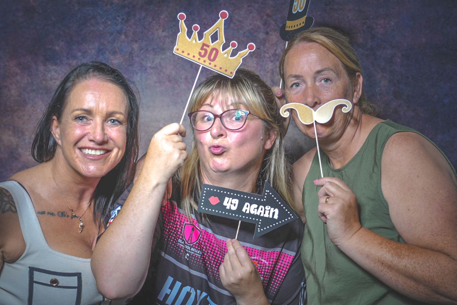 Three women posing with 50th birthday photo booth props including a crown and mustache at a party.