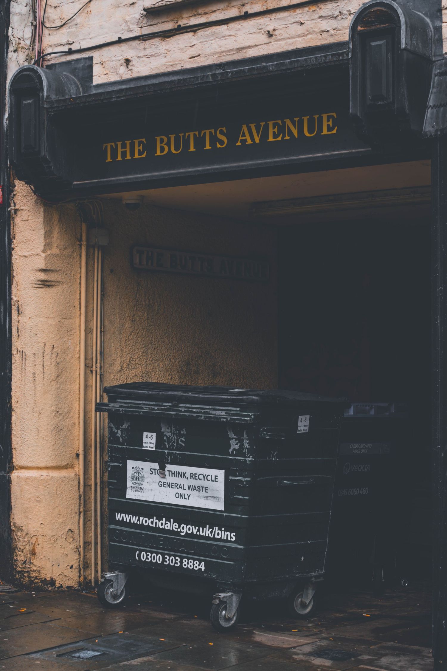 Dark storefront entrance with 'The Butts Avenue' signage and black waste bins visible against a beige wall.