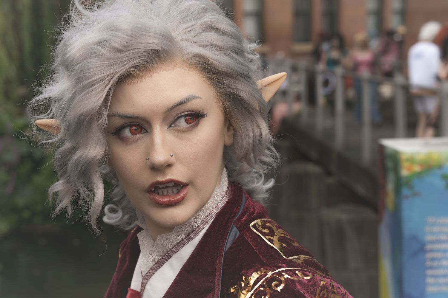Person with wavy silver hair wearing burgundy jacket and dark lipstick poses outdoors in urban setting.