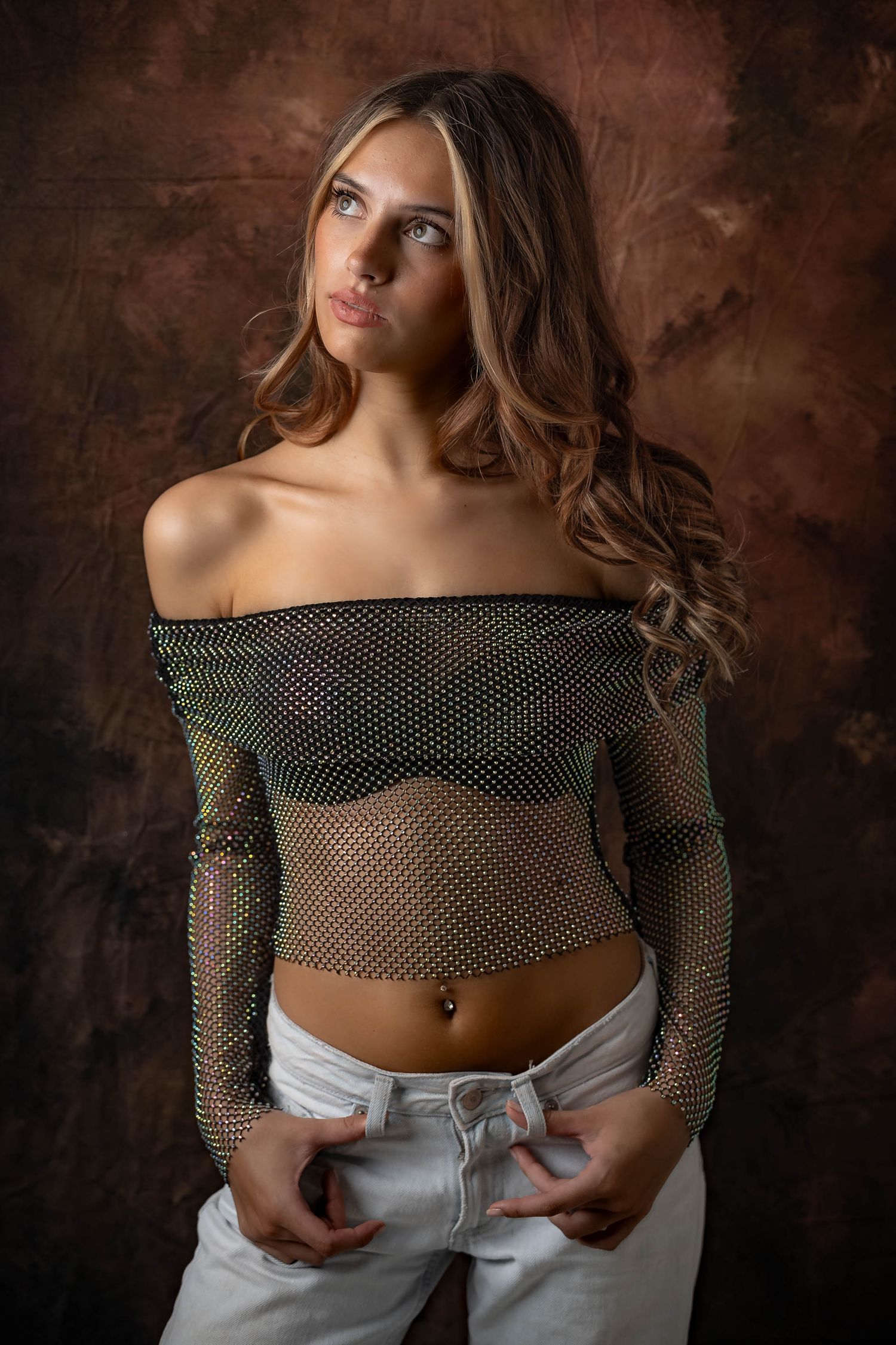 Young woman in sheer mesh off-shoulder top and white jeans posing against a dark brown textured studio background.