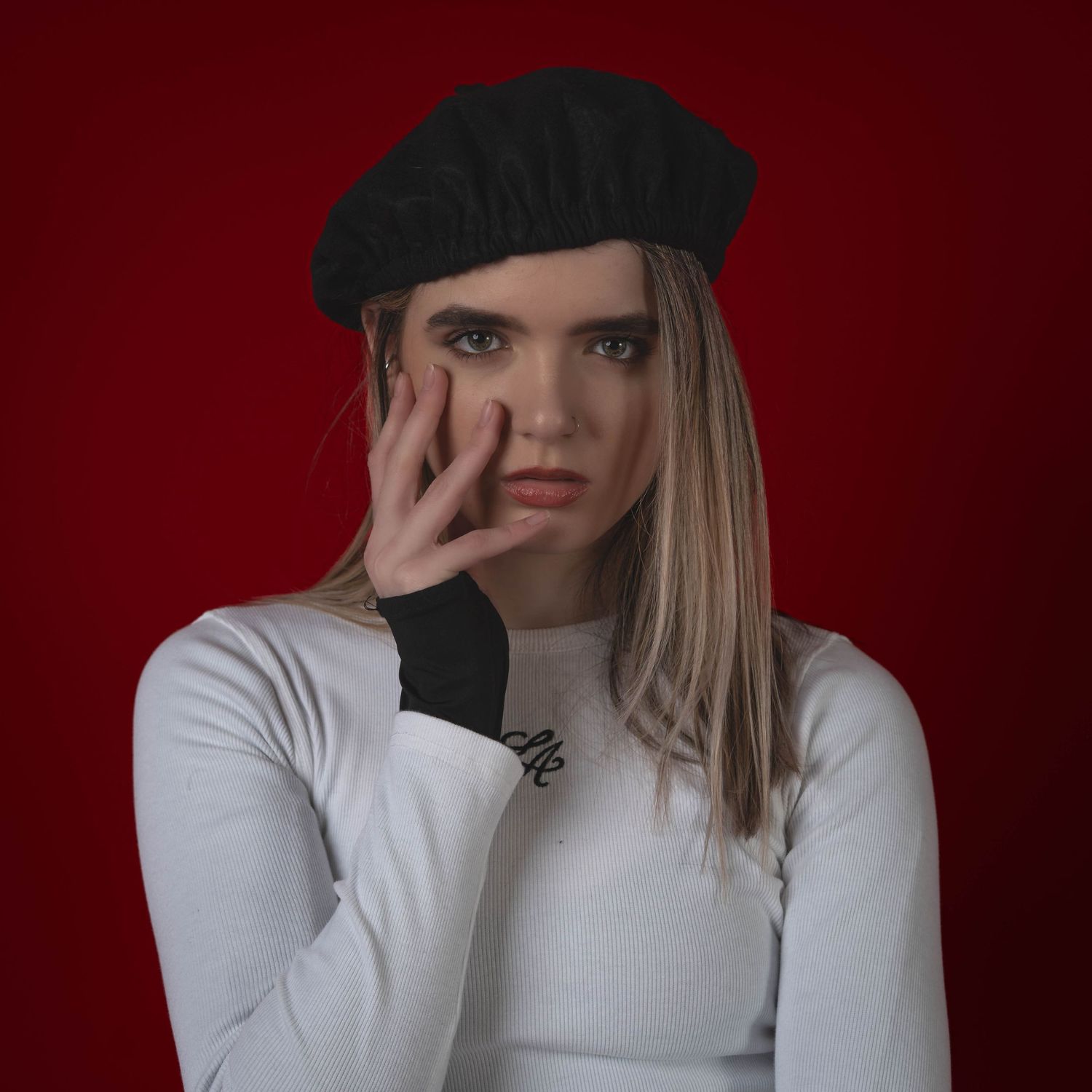 A person in a white top and black beanie poses against a dark red background in a creative portrait series.