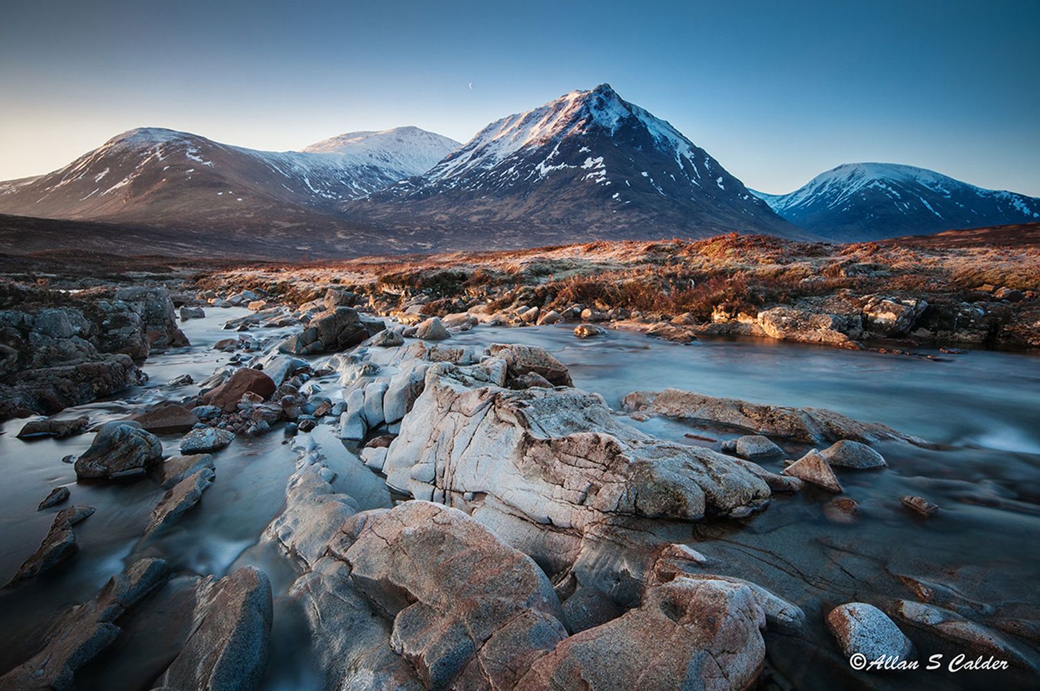 Dramatic panoramic view of snow-capped mountains and rugged terrain at sunset with golden light illuminating rocky landscape.