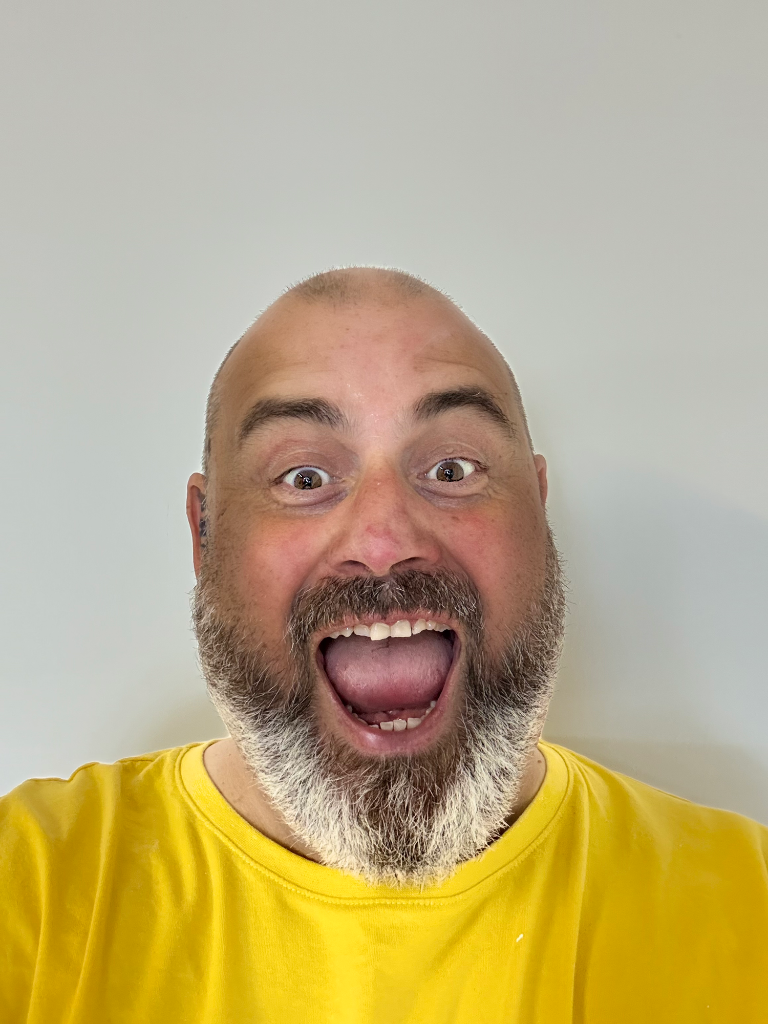 Bald man with gray beard wearing yellow shirt making excited open-mouth expression against white background.