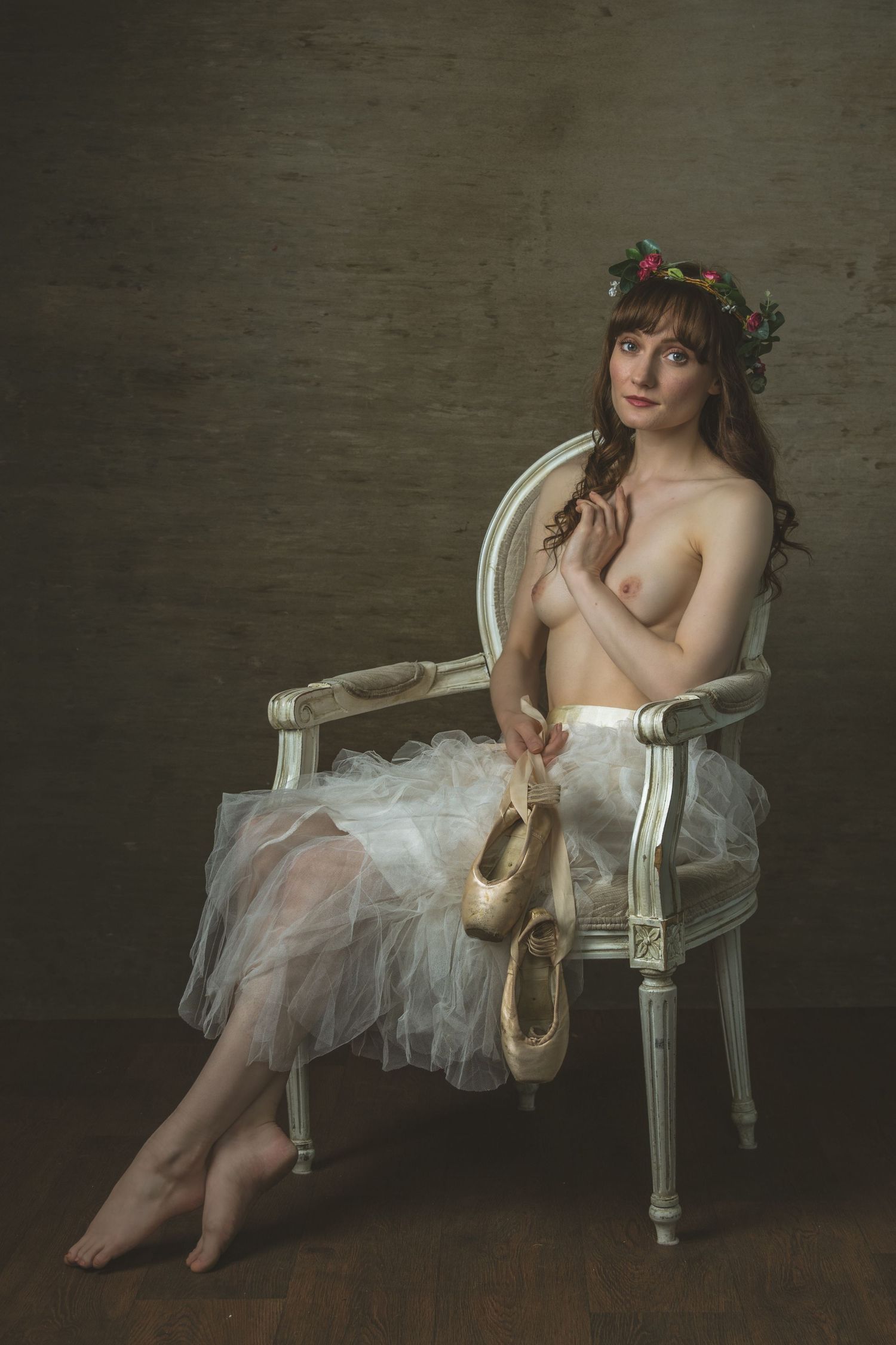 A ballerina wearing a floral crown and tutu skirt sits gracefully in an ornate chair holding pointe shoes.