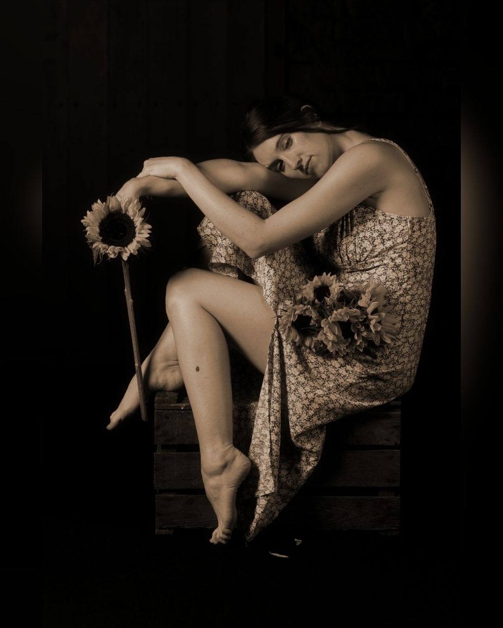 A woman in a floral dress sits in a pensive pose, her head resting on her hand in a contemplative expression.