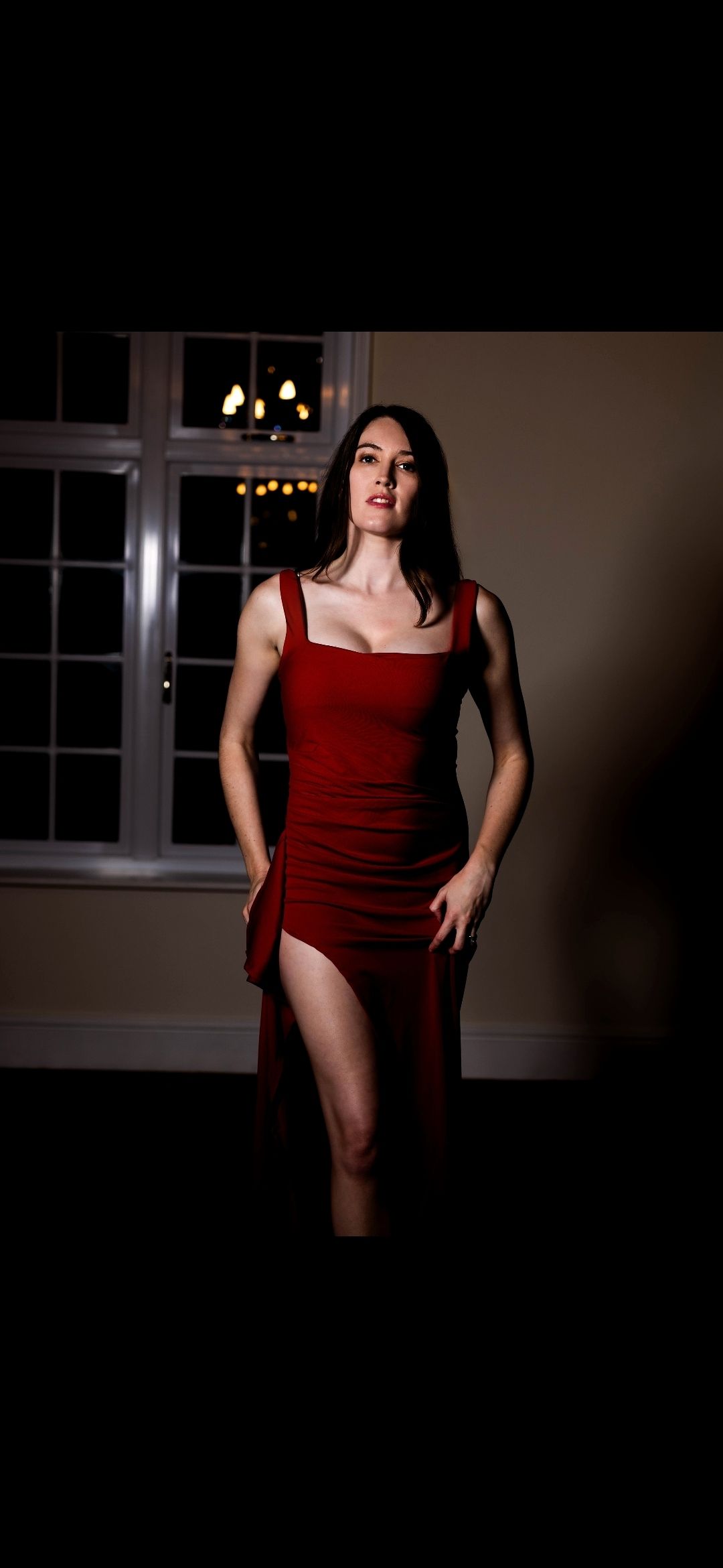 A woman in a red dress stands in a dimly lit room, her long dark hair framing her face.