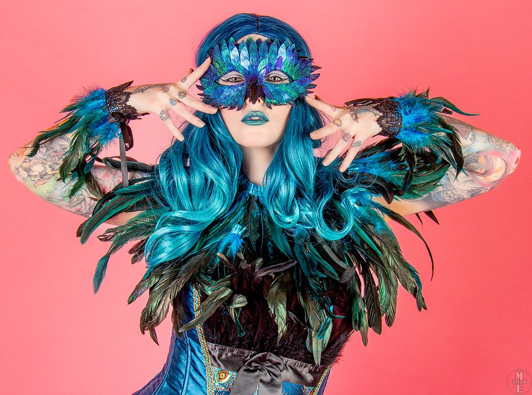 Woman in teal feathered costume and sequin masquerade mask with blue hair poses on pink background.