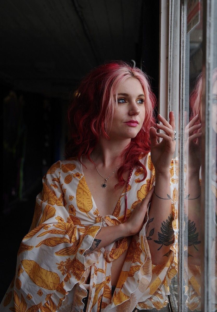 Woman with red hair in floral robe leaning against a window, tattoos visible, reflective glass background.