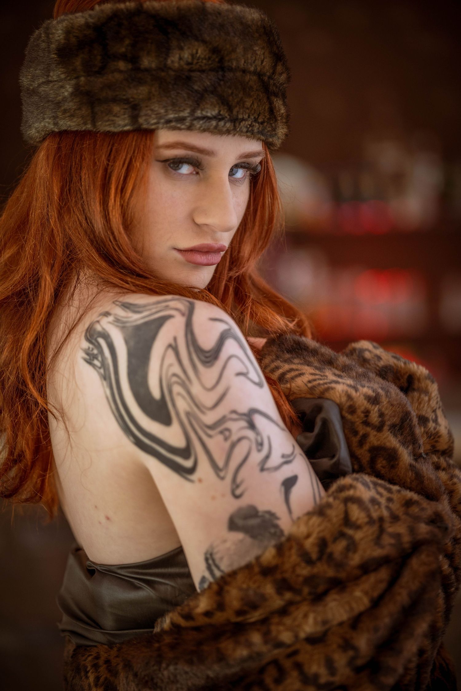 Red-haired woman with tattoos wearing a fur hat and leopard coat, posing elegantly in a moody indoor setting.