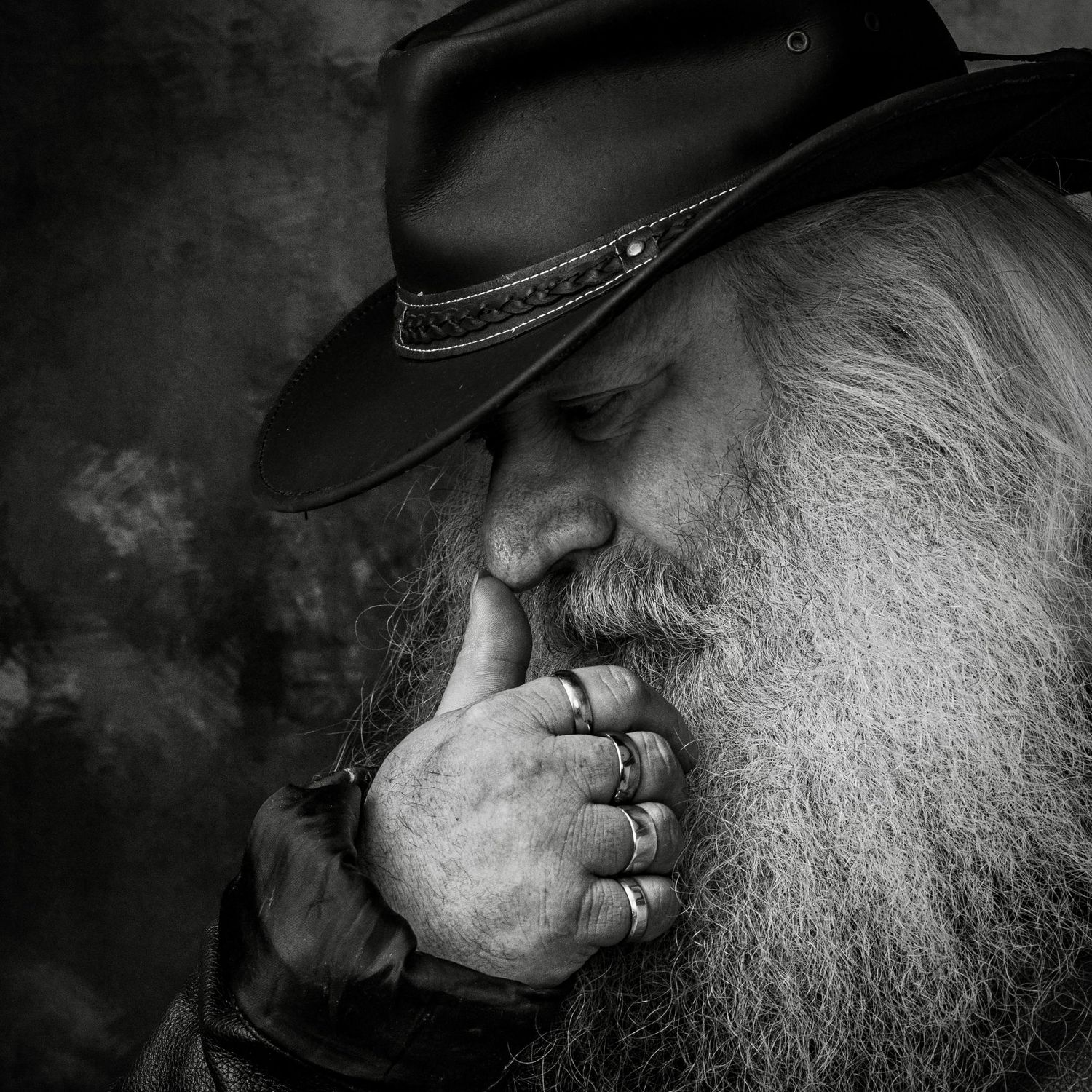 Black and white portrait of a bearded elderly man in a cowboy hat, hand raised thoughtfully to his lips, wearing rings.