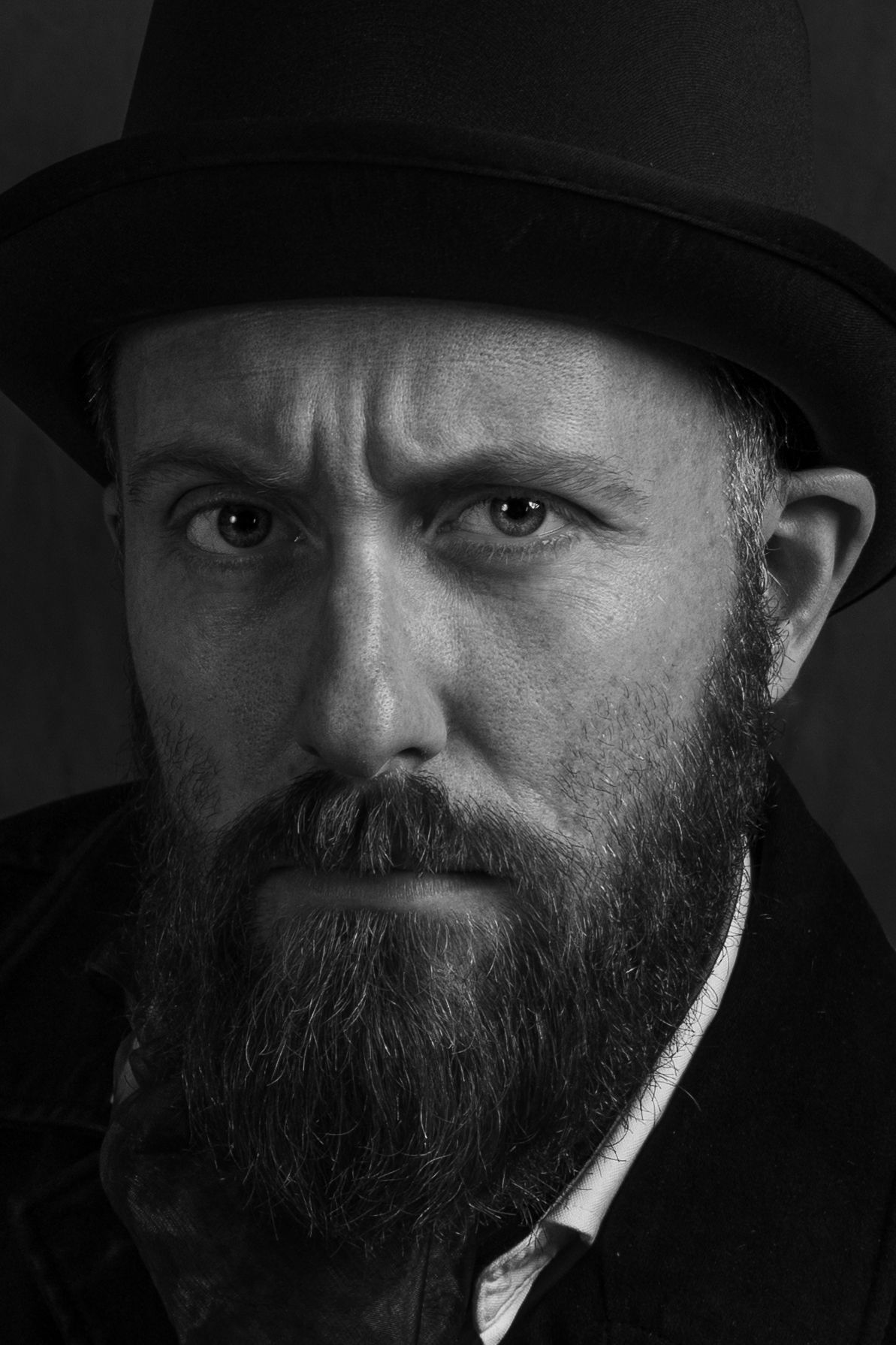 Black and white portrait of a bearded man wearing a dark hat, with an intense serious expression.